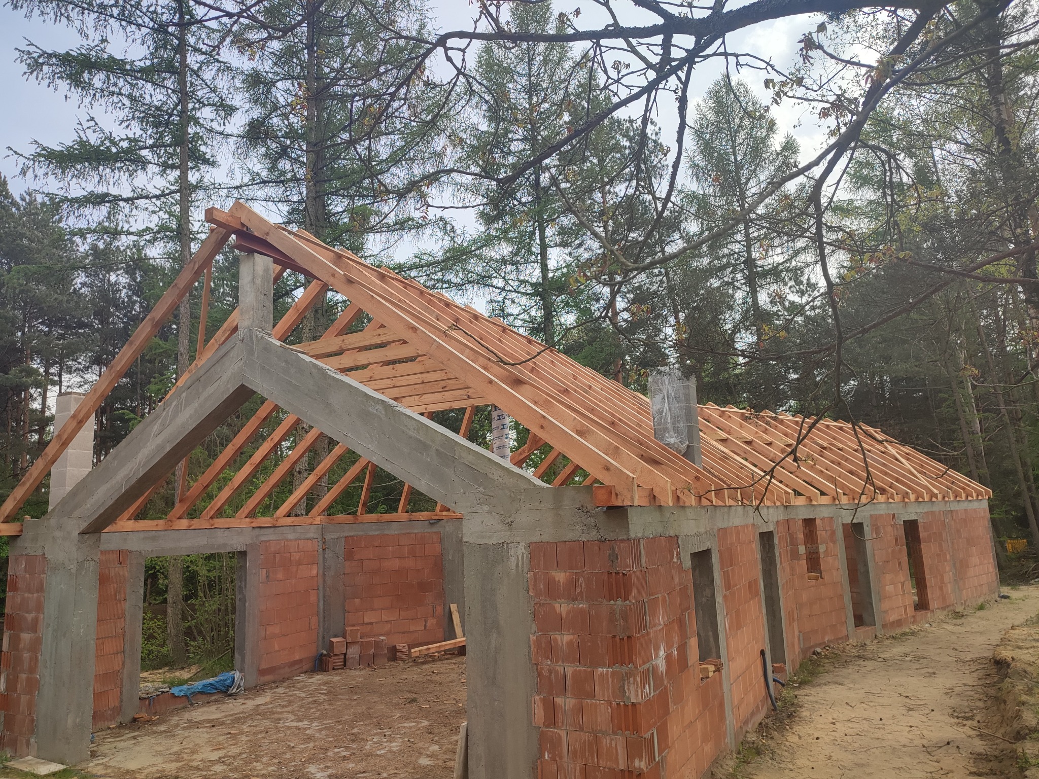 Surowy stan budynku z czerwonej cegły z drewnianą konstrukcją dachu na tle lasu. Widoczne niedokończone ściany i otwory okienne. Etap budowy domu jednorodzinnego.