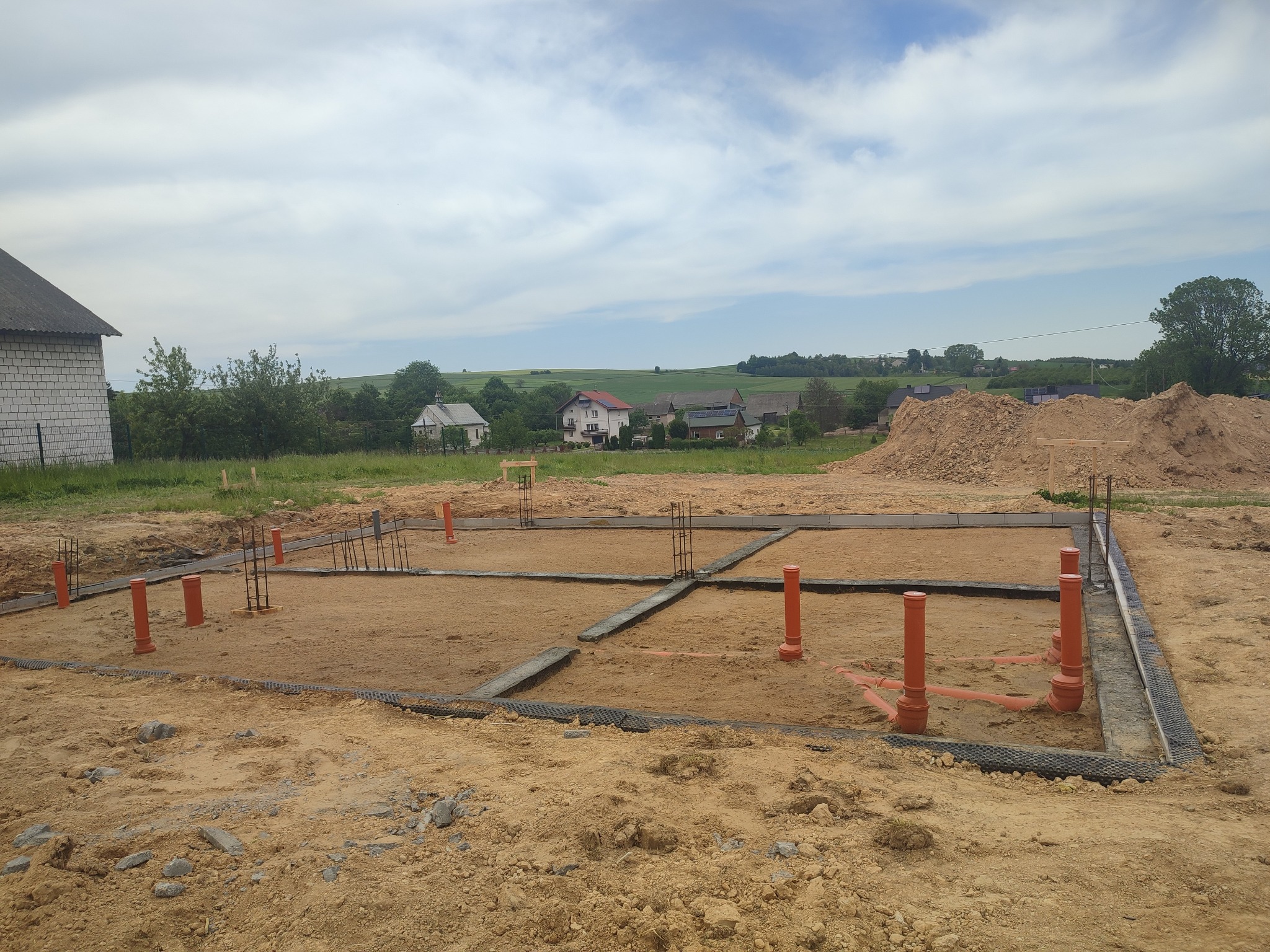 Fundamenty domu jednorodzinnego w trakcie budowy, widoczne zbrojenia i instalacje kanalizacyjne, w tle krajobraz wiejski z zabudowaniami.