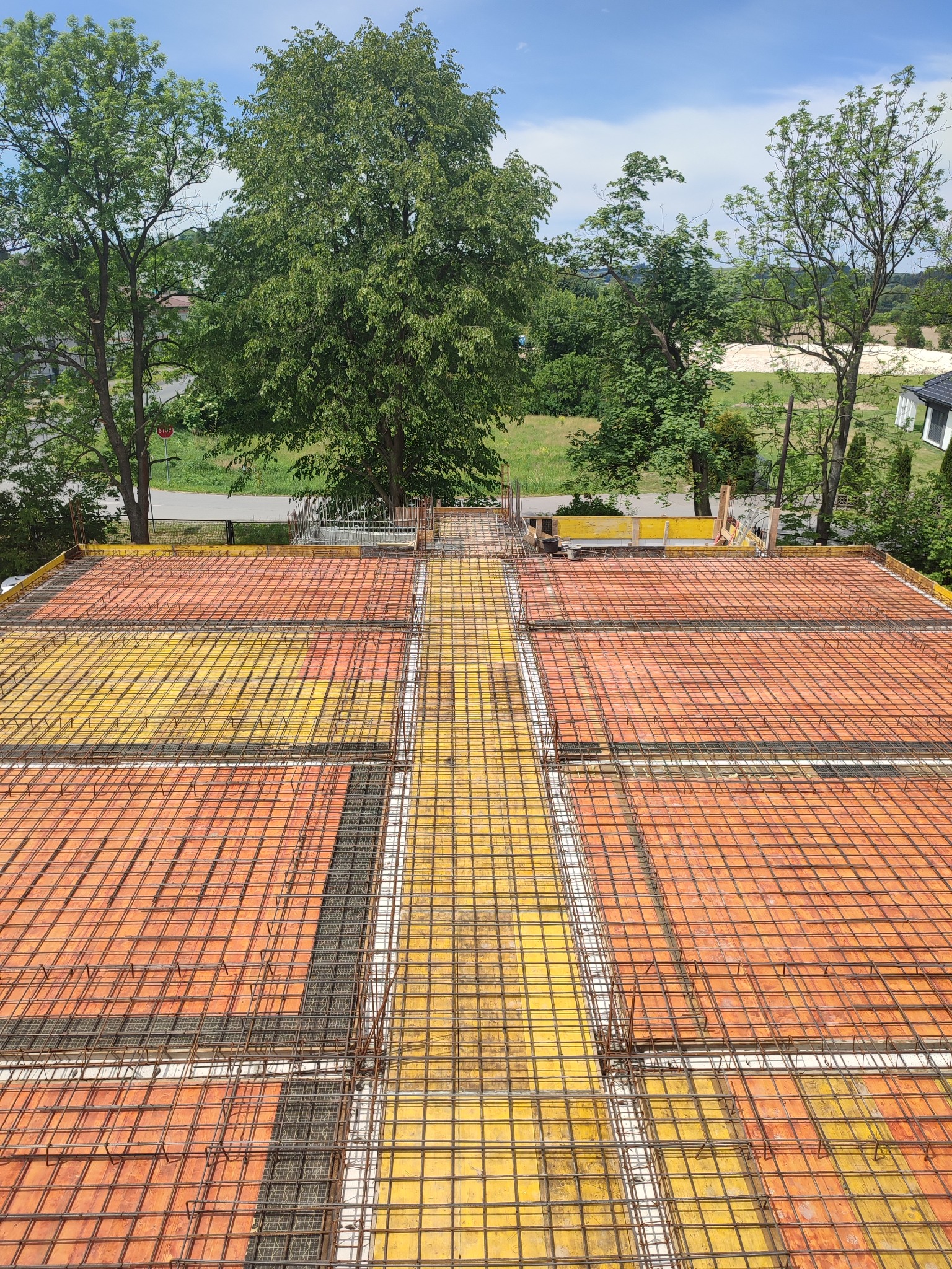 Zbrojenie stropu budynku z widoczną siatką i deskowaniem, przygotowane do zalania betonem. W tle zieleń drzew i fragment zabudowy.