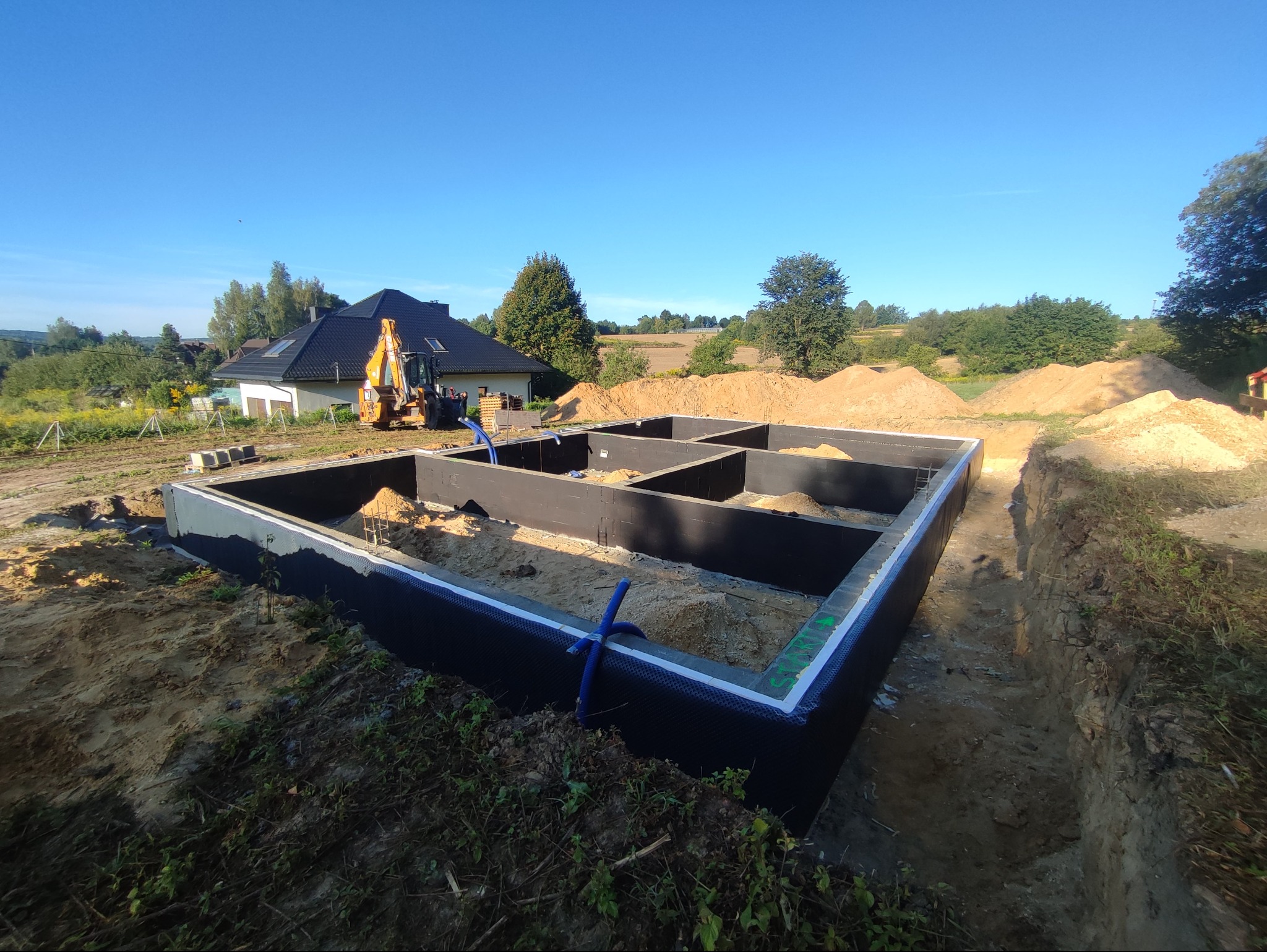 Zalane fundamenty domu jednorodzinnego z hydroizolacją, widoczne niebieskie rury, w tle koparka i dom w budowie, krajobraz wiejski, słoneczny dzień.