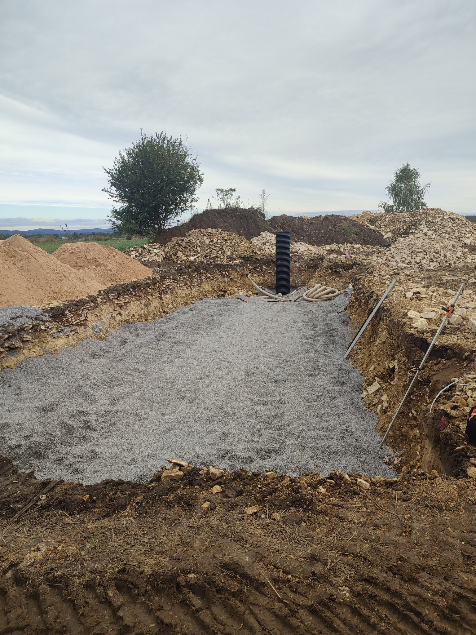 Przygotowany wykop pod fundamenty z wysypanym żwirem, widoczne rury i hałdy ziemi. Etap prac ziemnych przed wylaniem fundamentów pod budowę domu.