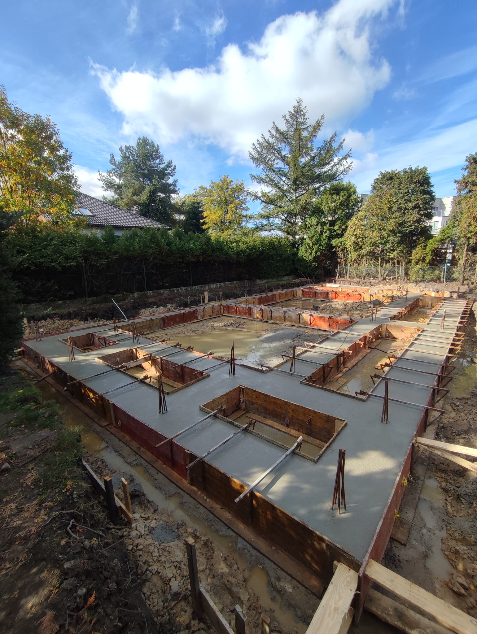 Zalane betonem fundamenty domu jednorodzinnego w drewnianym szalunku, wystające pręty zbrojeniowe, tło: zieleń drzew i błękitne niebo z chmurami.