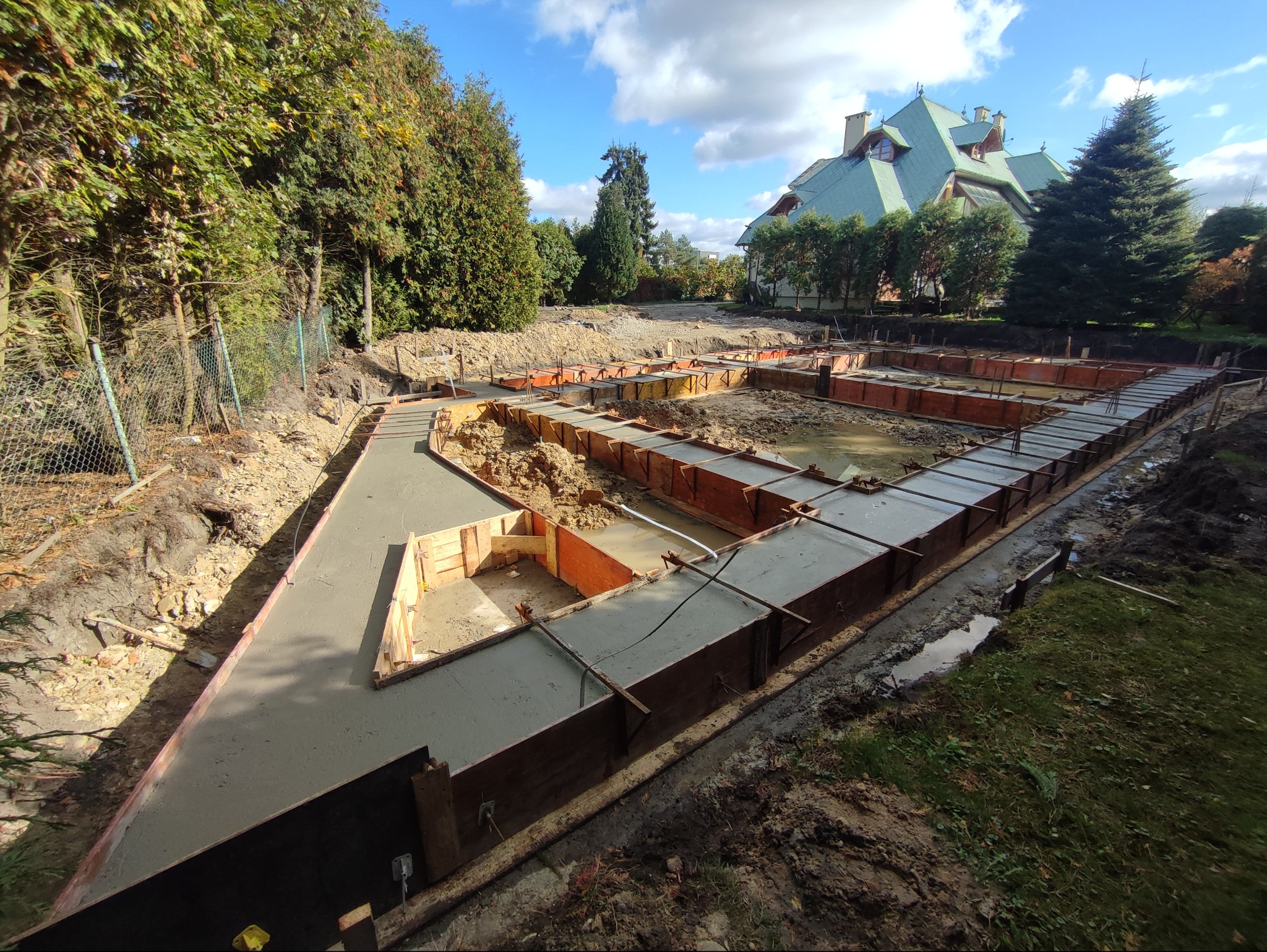 Wylane fundamenty pod budynek jednorodzinny z drewnianym szalunkiem, widok z góry na plac budowy w słoneczny dzień. W tle zieleń i budynek z zielonym dachem.