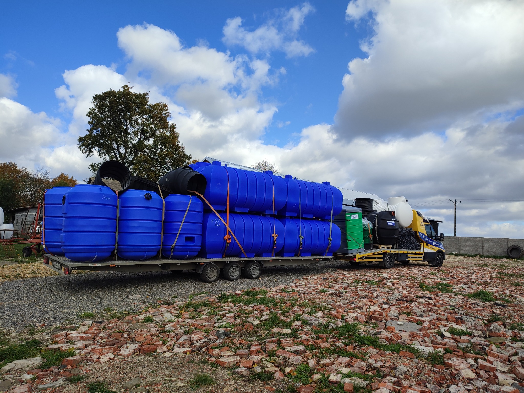 Ciężarówka z przyczepą przewozi niebieskie zbiorniki na tle błękitnego nieba z chmurami. Na pierwszym planie gruz ceglany i trawa.
