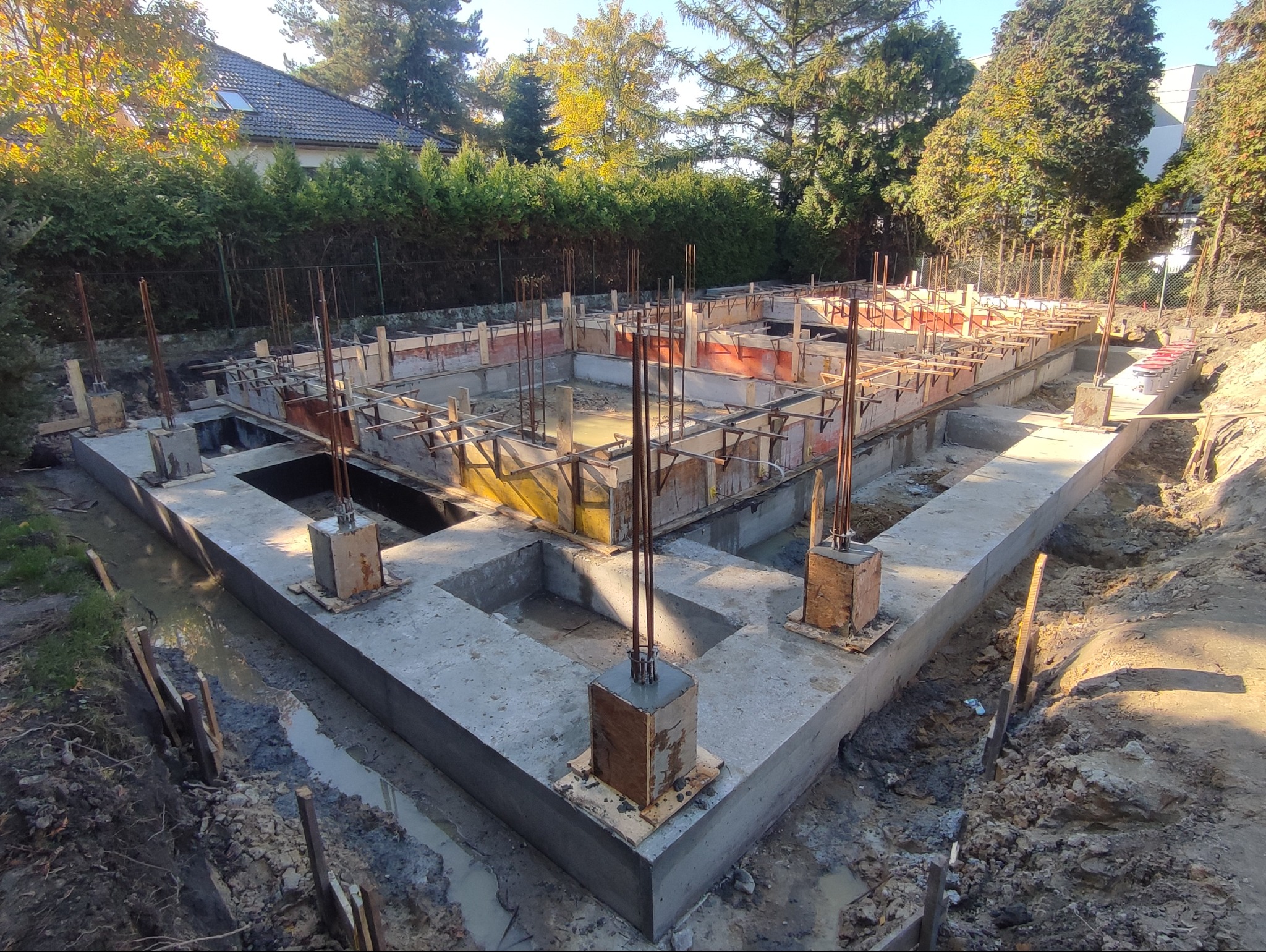 Zalane fundamenty domu jednorodzinnego z widocznym szalunkiem i wystającymi prętami zbrojeniowymi. Prace murarskie w toku, teren budowy.