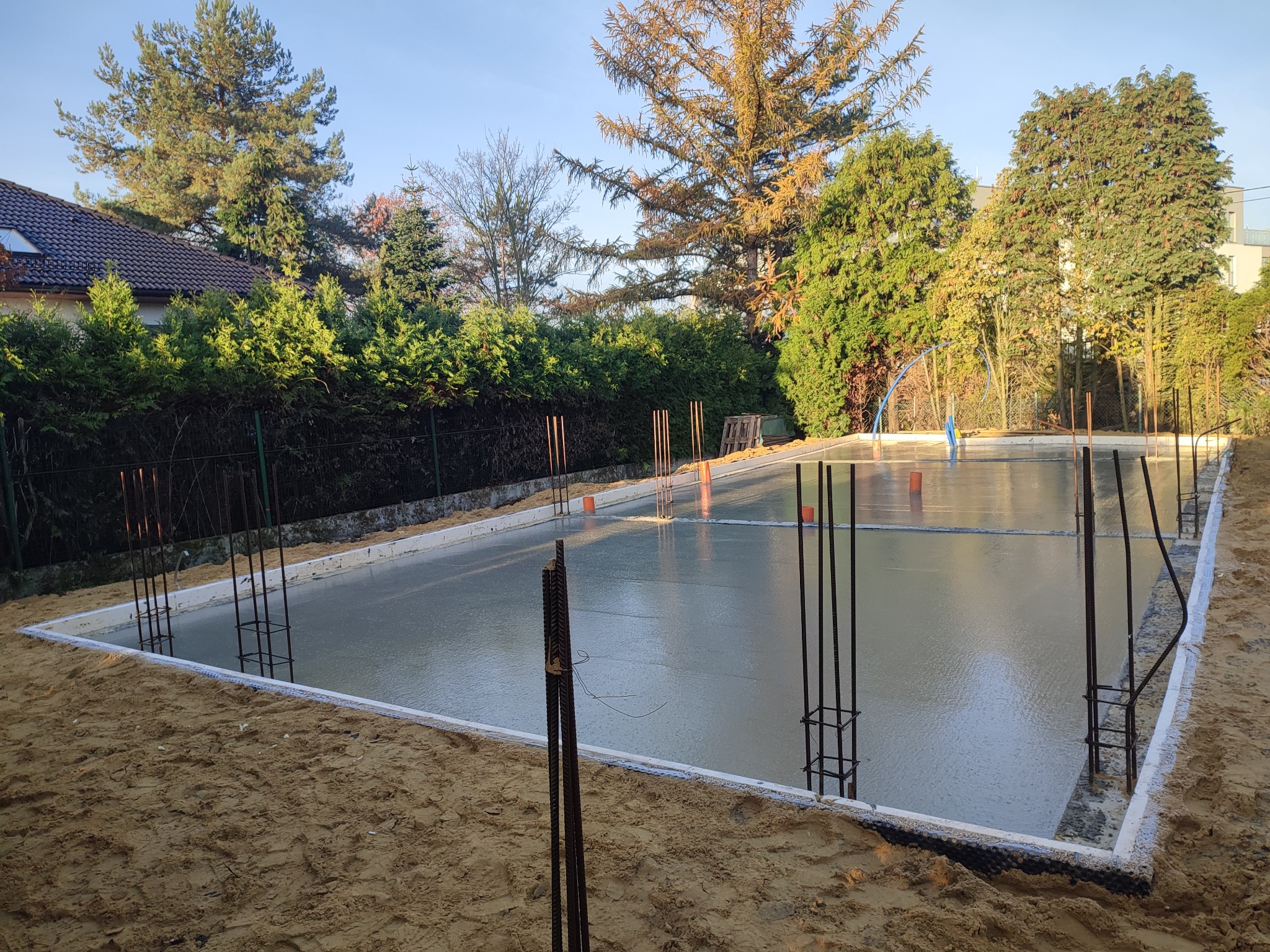 Zalana betonem płyta fundamentowa z wystającymi prętami zbrojeniowymi, otoczona piaskiem i zielenią. Widok na wczesny etap budowy domu.