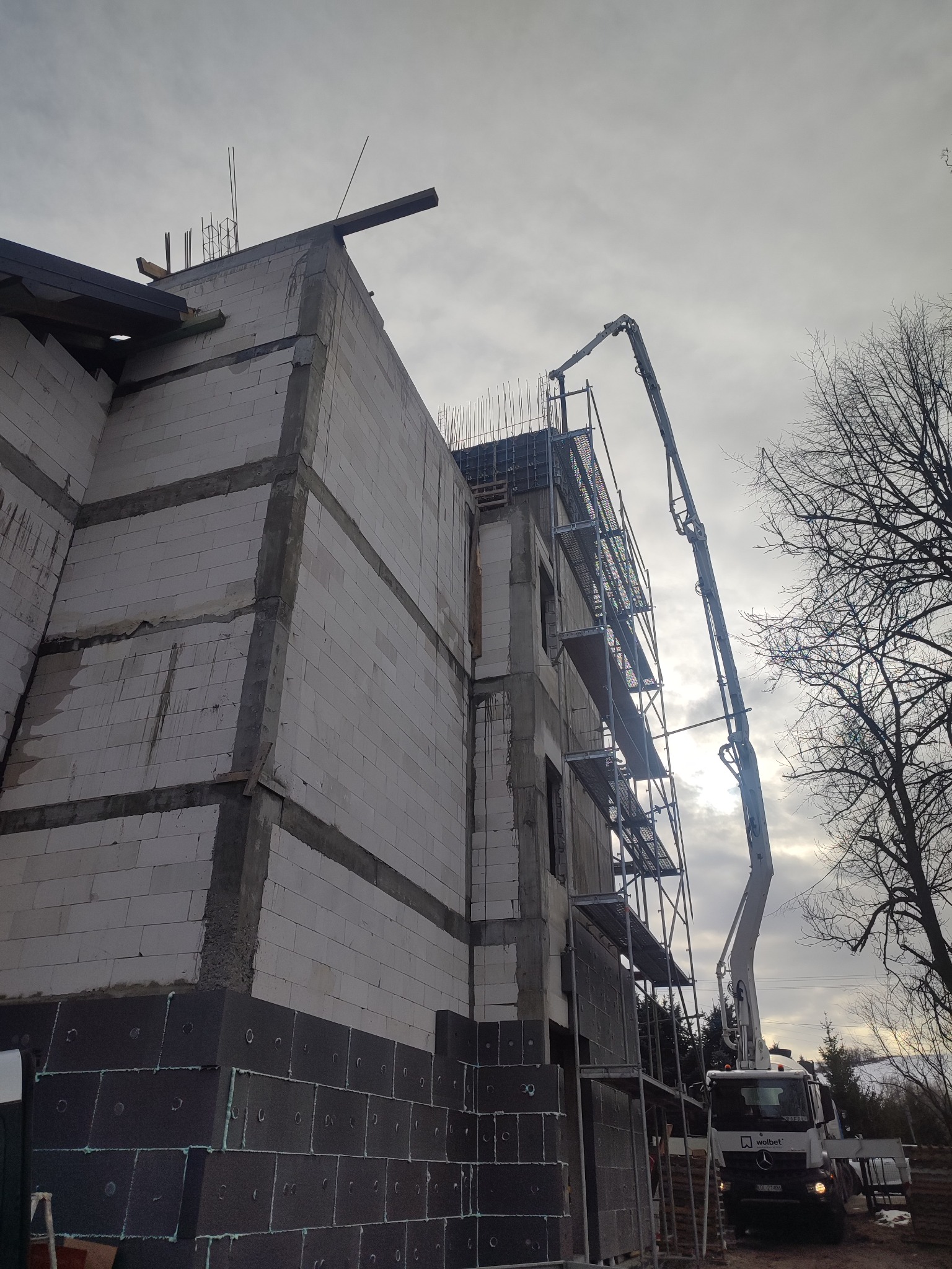 Budowa domu z białych bloczków, widoczne rusztowania i pompa do betonu na tle pochmurnego nieba. Etap wznoszenia ścian, zbrojenia i wylewania betonu.