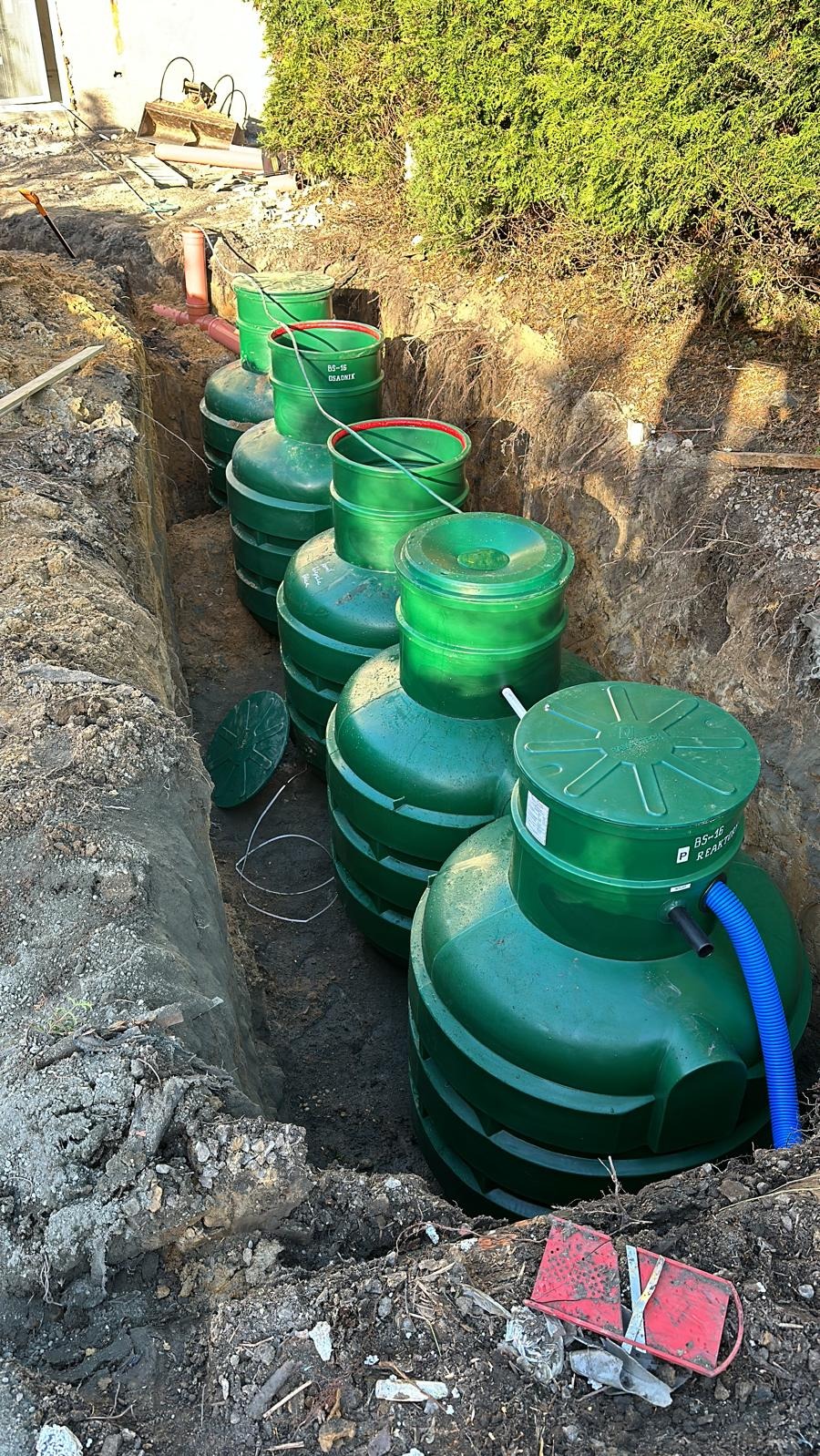 Szereg zielonych zbiorników oczyszczalni ścieków w wykopie, widoczne rury i elementy instalacji. Częściowo zasypana ziemią, w tle zieleń. Widok z góry.