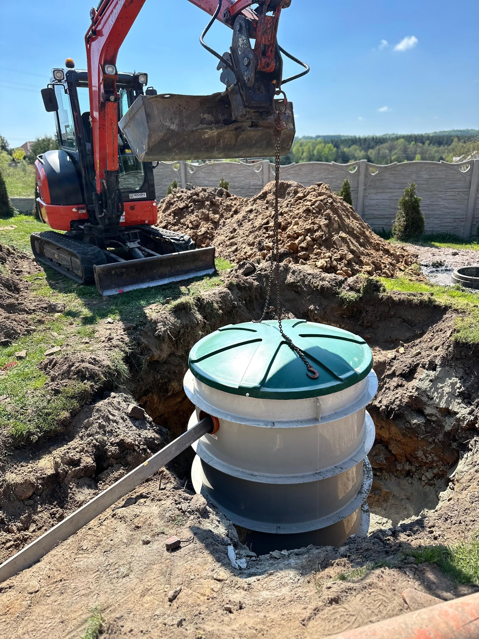 Montaż przydomowej oczyszczalni ścieków: biały zbiornik z zieloną pokrywą opuszczany do wykopu za pomocą koparki na tle ogrodzenia i zieleni.