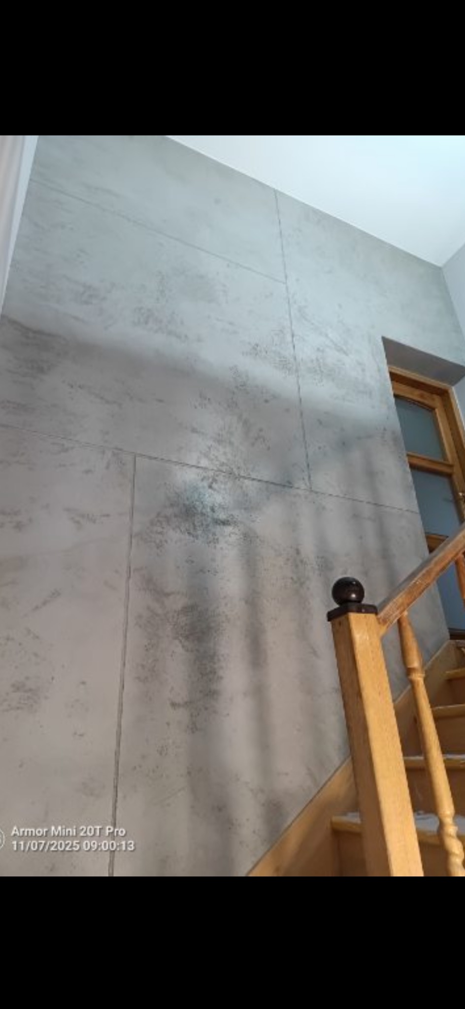 Ściana z efektem betonu architektonicznego, podzielona na panele, widoczna klatka schodowa z drewnianą balustradą i fragment drzwi. Wykończenie wnętrza w szarościach.