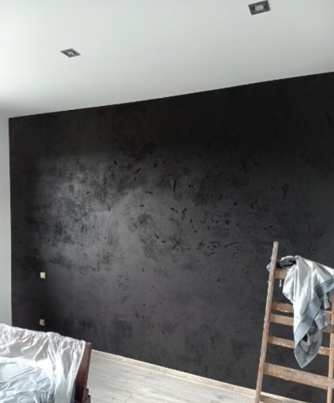 Ściana z efektem dekoracyjnym w odcieniach szarości i czerni, widoczna drabina malarska i fragment łóżka. Minimalistyczne oświetlenie sufitowe.