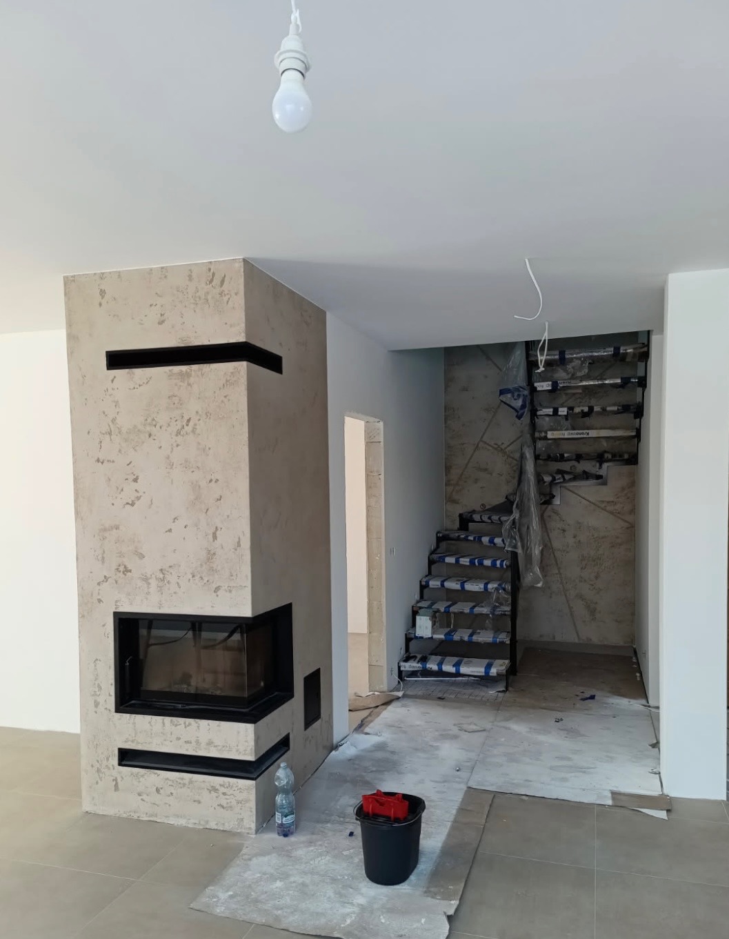 Nowoczesny kominek z dekoracyjną strukturą obok schodów w trakcie wykończenia. Widoczne narzędzia i materiały budowlane. Minimalistyczny design.