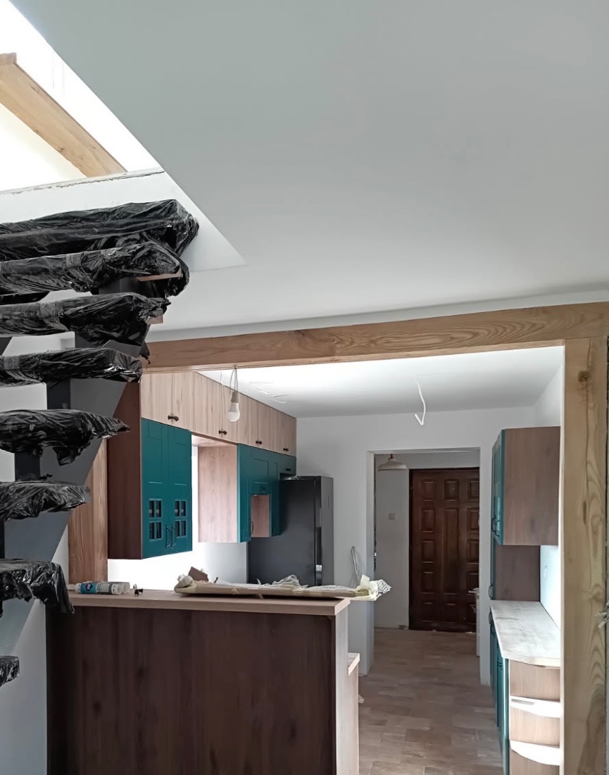 Wnętrze w trakcie wykańczania: zabudowa kuchenna z turkusowymi akcentami, drewniane elementy konstrukcyjne i schody zabezpieczone folią ochronną. Widok na korytarz.