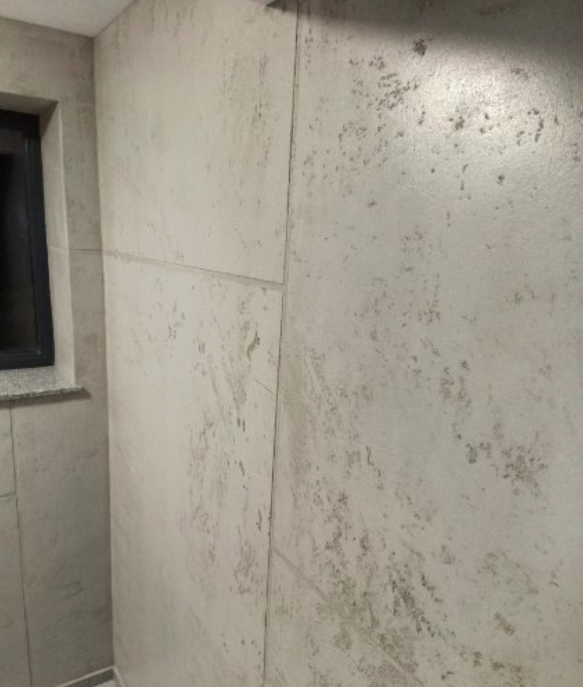 Ściana wykończona w odcieniach szarości z efektem dekoracyjnym, imitującym kamień lub beton. Widoczne łączenia płyt i fragment okna z ciemną ramą.