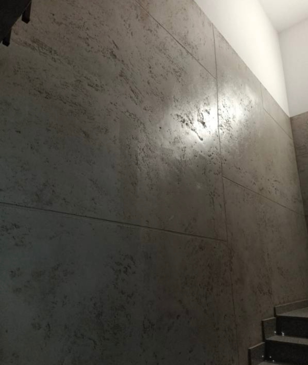 Dekoracyjna ściana w odcieniach szarości z efektem betonu architektonicznego, widoczne łączenia płyt. U dołu fragment schodów.