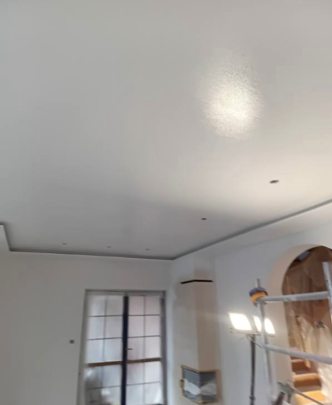 Białe, świeżo pomalowane wnętrze z oknem i dekoracyjnym kominkiem. Widoczny fragment rusztowania i lampa oświetlająca sufit. Minimalistyczny design.