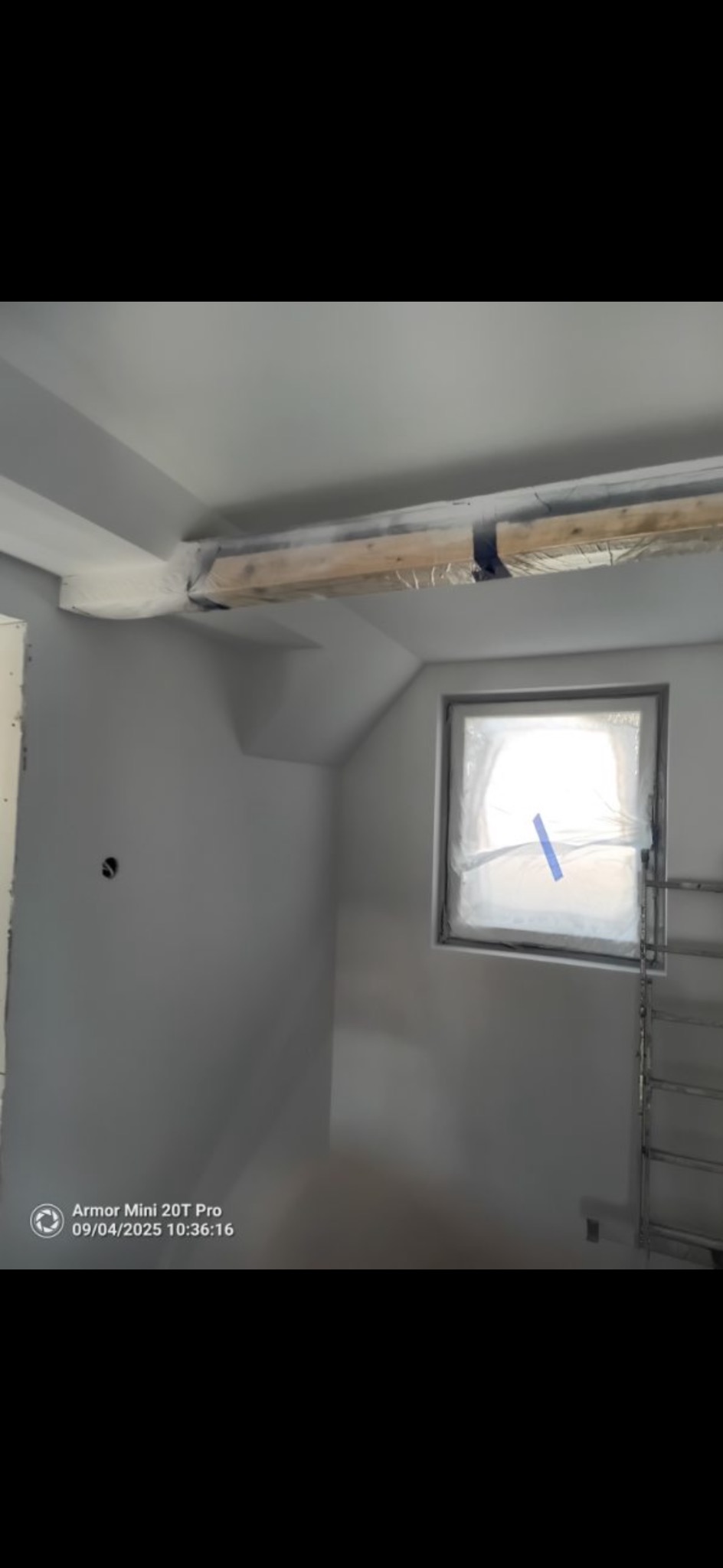 Pomieszczenie po malowaniu na szaro, z zabezpieczonym oknem folią i drewnianą belką pod sufitem. Widoczna drabina. Świeżo pomalowane ściany i sufit.