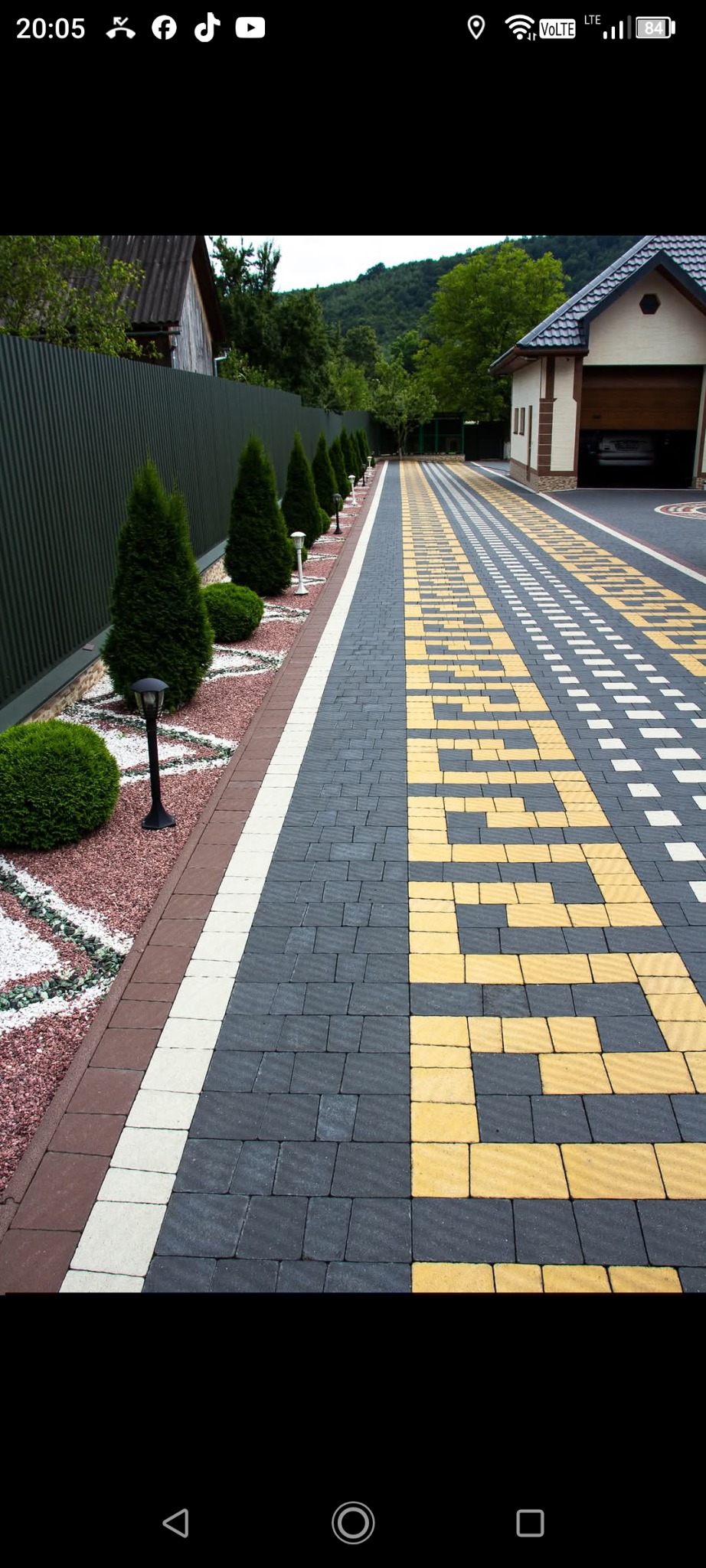 Ogród z geometrycznym wzorem z kostki brukowej w odcieniach szarości i żółci, obok żywopłot z tujami i lampami. W tle garaż i zieleń.