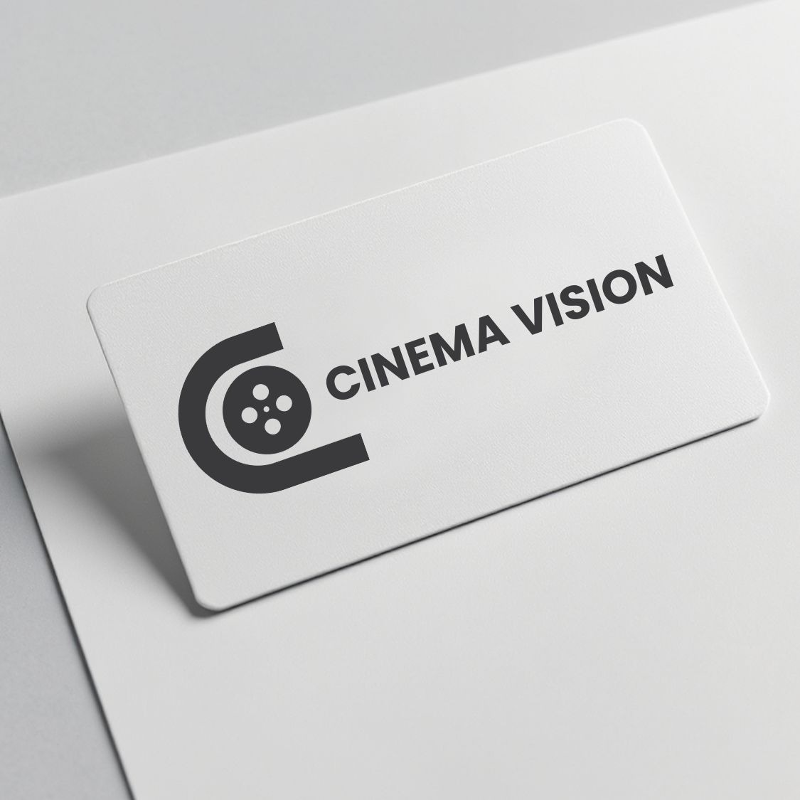 Minimalistyczny, czarno-biały projekt logo 'Cinema Vision' z symbolem filmowej taśmy na wizytówce, leżącej na jasnym tle. Prosty, nowoczesny design.