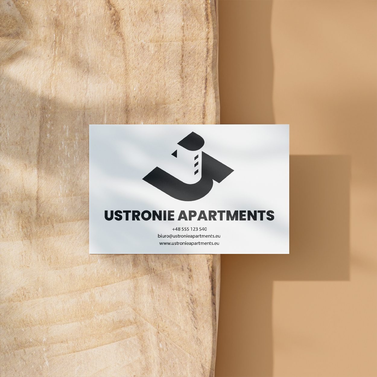 Wizytówka Ustronie Apartments z czarnym logo na białym tle, leżąca na drewnianej powierzchni z cieniem. Minimalistyczny design, wyraźne dane kontaktowe.
