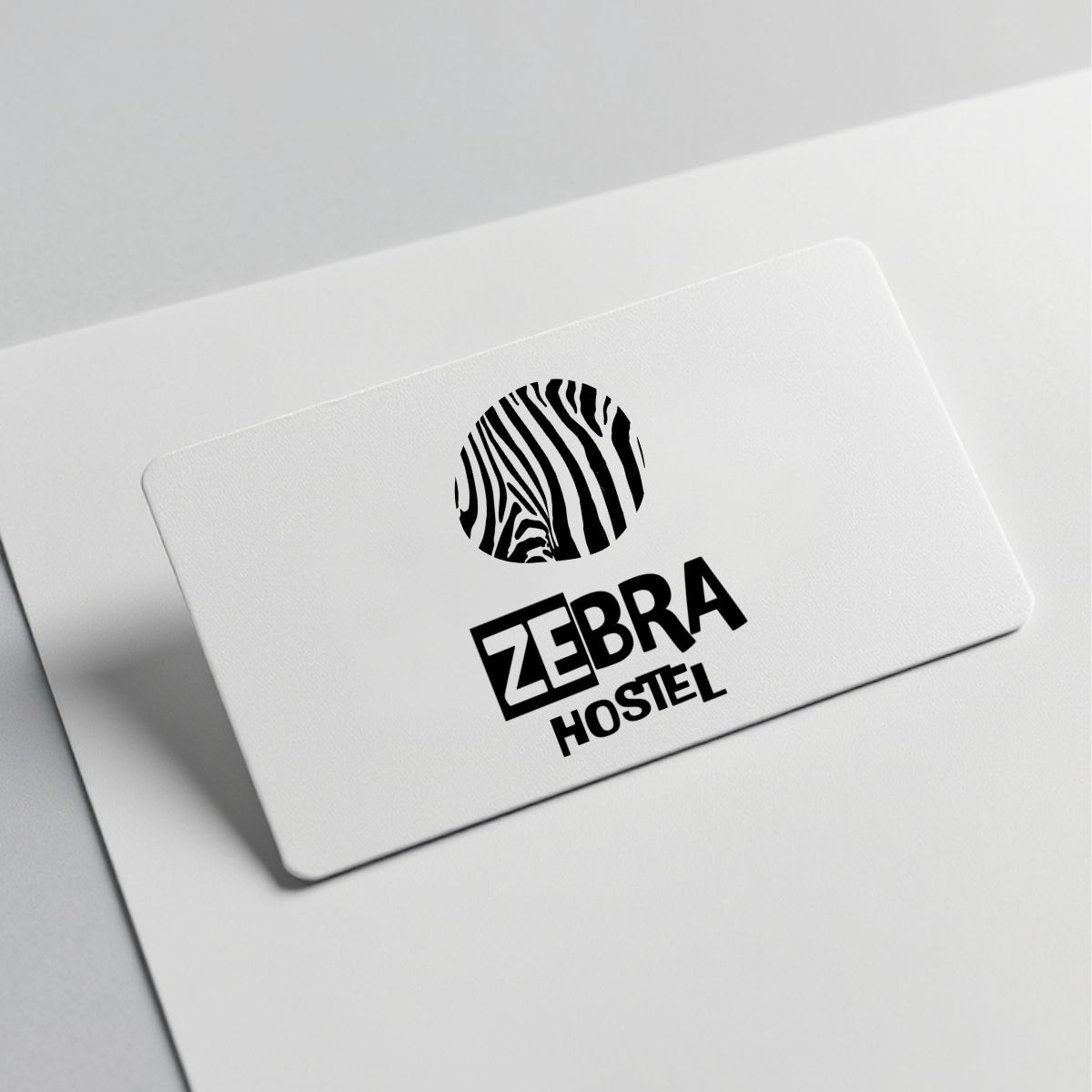 Minimalistyczny projekt logo 'Zebra Hostel' na białej wizytówce, z okrągłym symbolem zebry w paski. Prosty, nowoczesny design na jasnym tle.