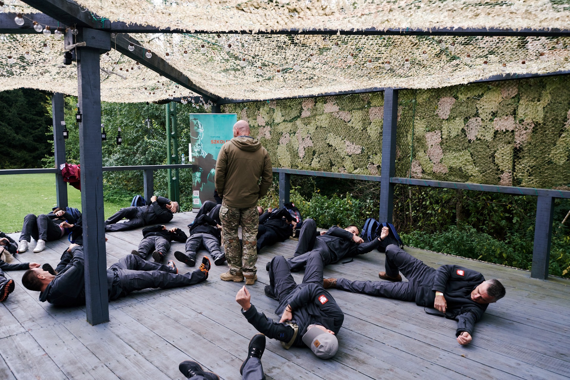 Instruktor nadzoruje ćwiczenia grupy na drewnianym podeście. Uczestnicy w szarych kombinezonach leżą na plecach. W tle zieleń i baner szkoleniowy.