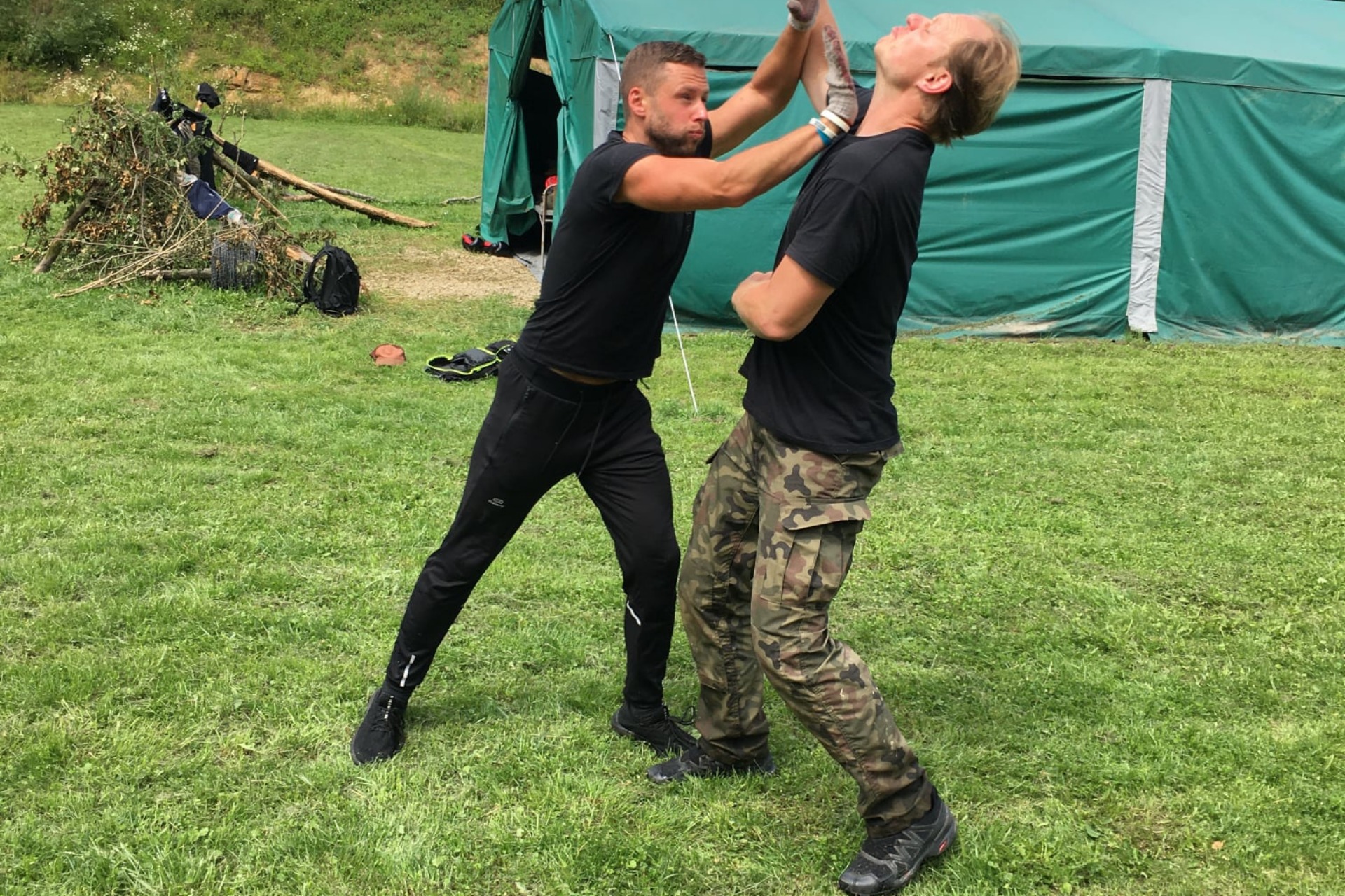 Instruktor demonstruje technikę samoobrony na kursie, blokując atak na szyję. Widoczny zielony namiot w tle i trawiasta przestrzeń.