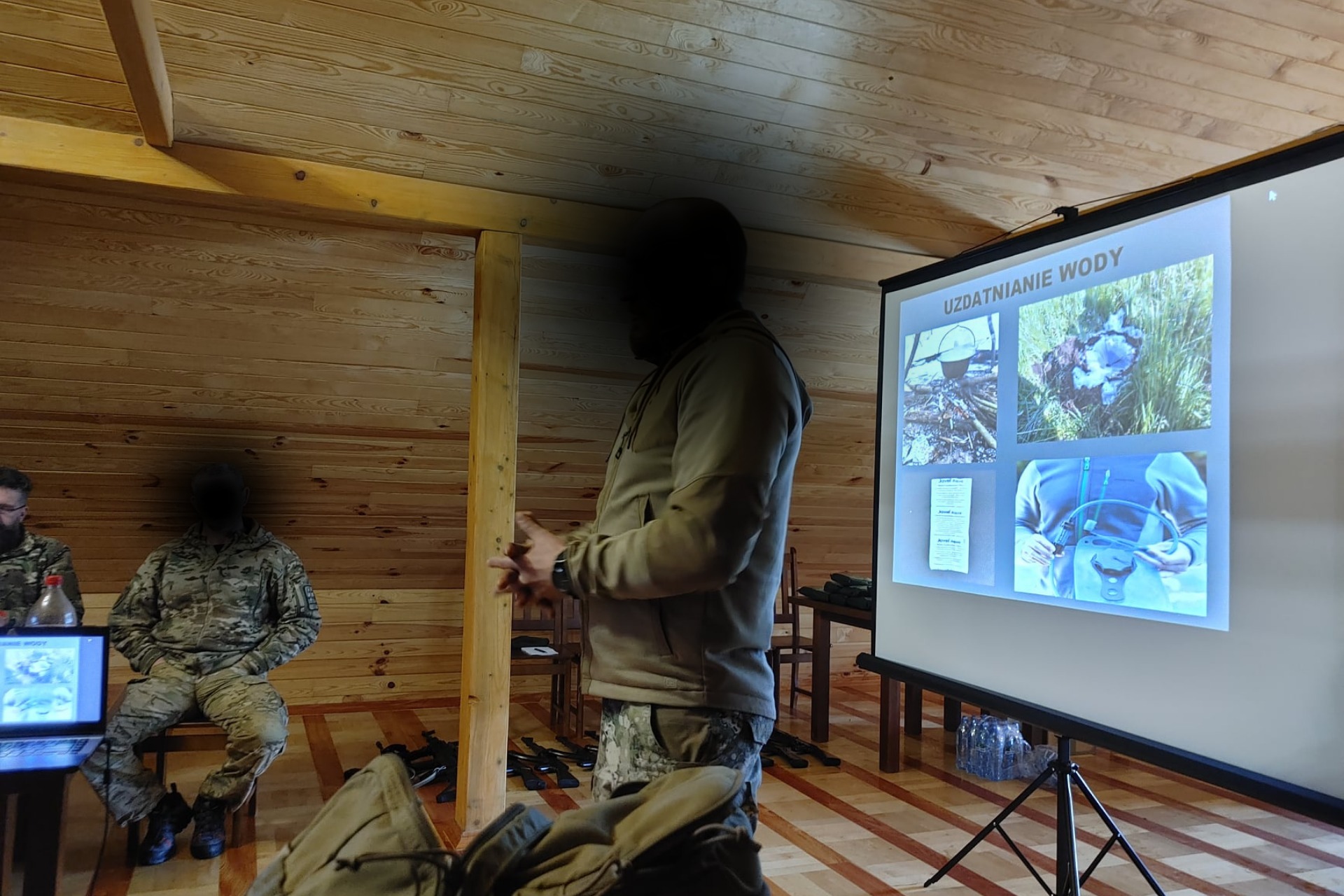 Prezentacja o uzdatnianiu wody podczas szkolenia technicznego w drewnianym wnętrzu. Projektor wyświetla zdjęcia, a uczestnicy w strojach taktycznych słuchają prelegenta.