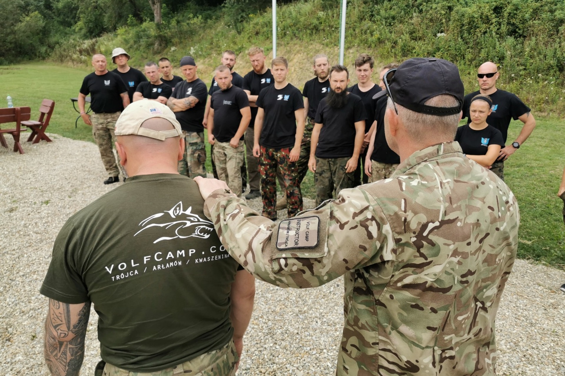 Instruktor w kamuflażu prowadzi grupę w czarnych koszulkach z logo Wolfcamp na zewnątrz. Uczestnicy w różnorodnych spodniach, teren trawiasty w tle.