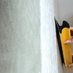 BUILDERSCRAFT Sp. z o.o. - Mężczyzna z brodą, z profilu, wygładza ścianę żółtą szpachelką. Ściana pokryta świeżą warstwą gładzi, widoczna faktura. Oświetlenie naturalne, jasne.