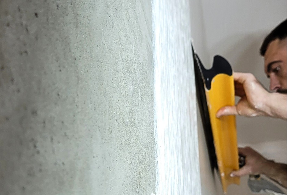 Mężczyzna z brodą, z profilu, wygładza ścianę żółtą szpachelką. Ściana pokryta świeżą warstwą gładzi, widoczna faktura. Oświetlenie naturalne, jasne.