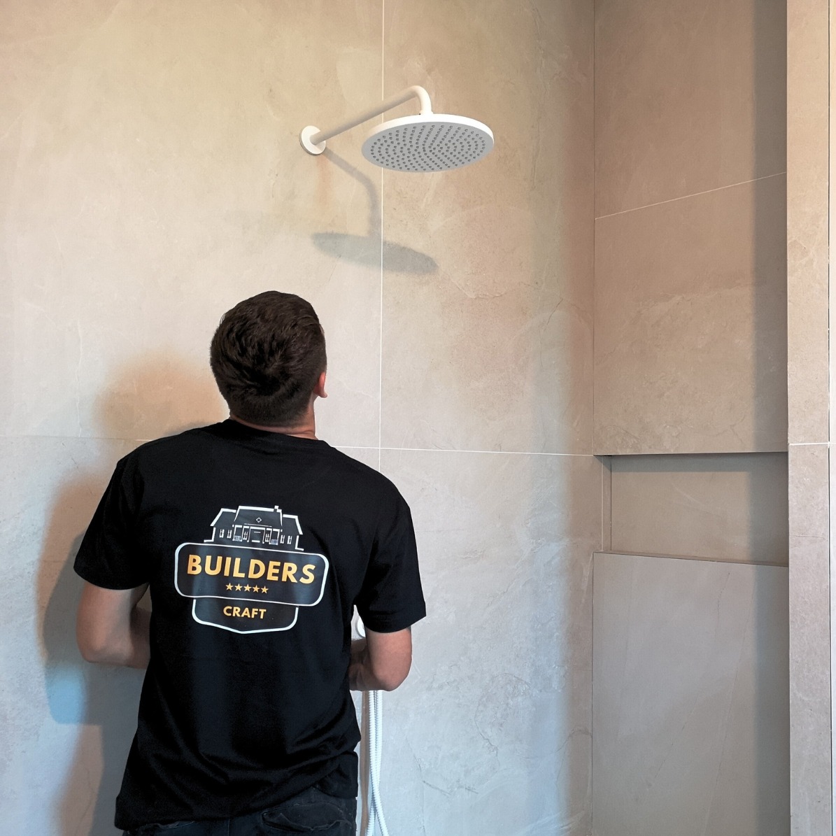 Instalacja deszczownicy w beżowej łazience z wnęką. Mężczyzna w czarnej koszulce z logo firmy instaluje białą deszczownicę na tle płytek imitujących kamień.