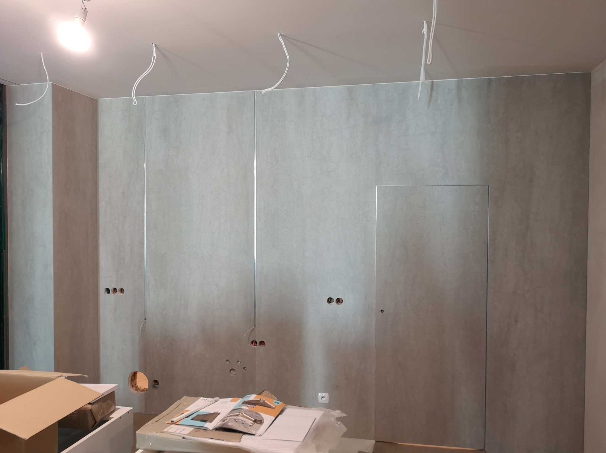 Ściana wykończona panelami imitującymi beton, widoczne otwory na gniazdka i kable, sufit z wystającymi przewodami, kartony i katalogi na podłodze. Wnętrze w trakcie wykańczania.