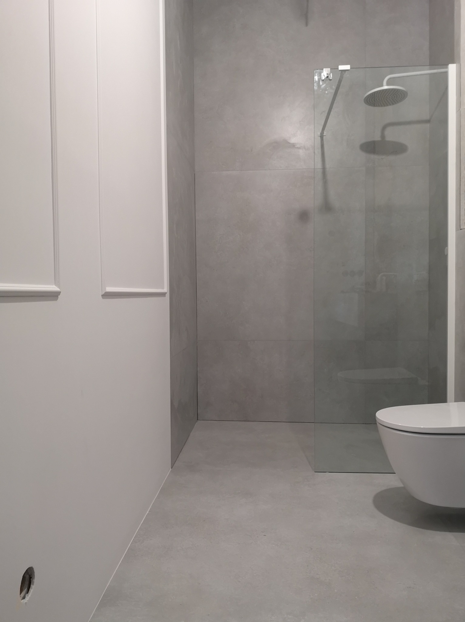 Minimalistyczna łazienka w szarościach z kabiną prysznicową typu walk-in i podwieszaną toaletą. Ściany wykończone płytami imitującymi beton, białe listwy dekoracyjne.