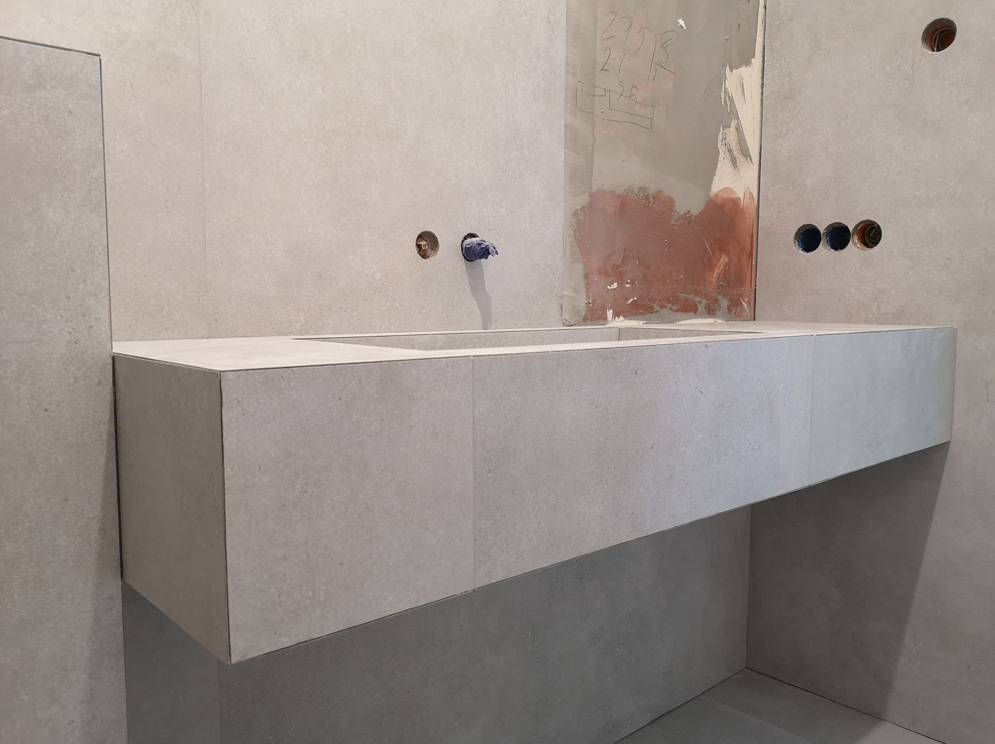 Wykończona, minimalistyczna umywalka z szarej płyty w łazience. Widoczne otwory na armaturę i instalację elektryczną. Ściany i podłoga również wyłożone płytami.