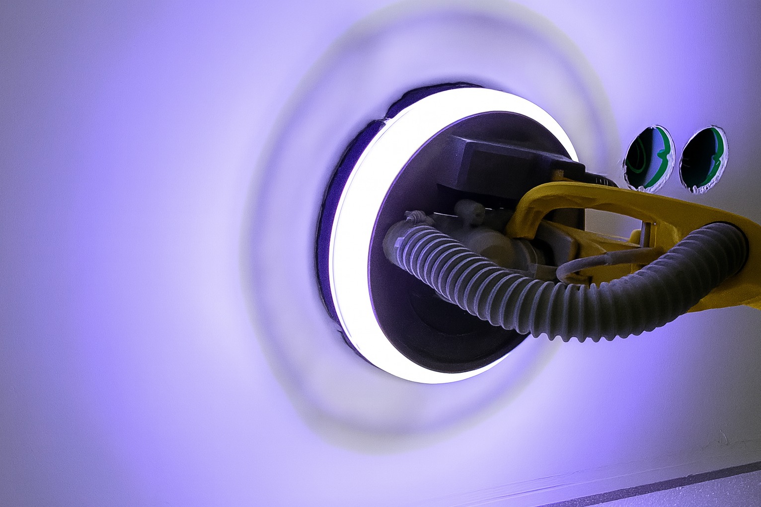 Szlifierka z oświetleniem LED wyrównuje białą ścianę, obok widoczne otwory na gniazdka. Szlifowanie bezpyłowe z odkurzaczem. Fioletowe oświetlenie.