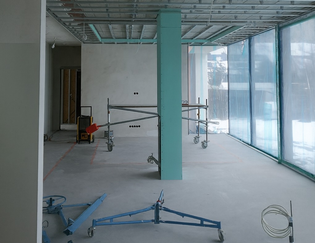 Wnętrze w trakcie prac wykończeniowych: metalowa konstrukcja sufitu, rusztowania, kolumna z płyty gipsowej, folia ochronna na oknach, narzędzia do montażu płyt.