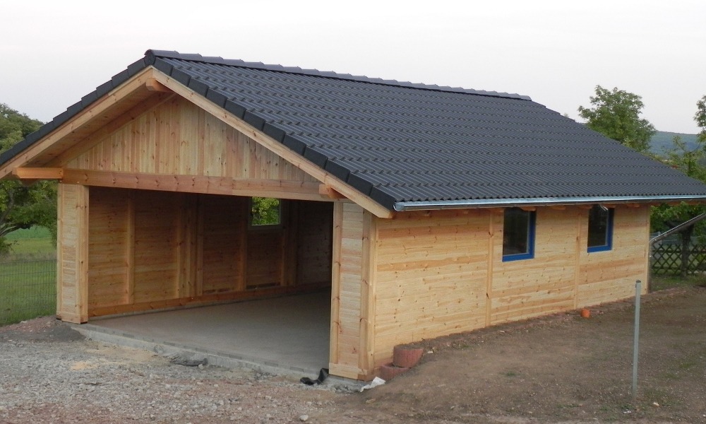 Drewniany garaż z ciemną dachówką falistą i betonową wylewką, widok z zewnątrz, w tle zieleń. Dwa okna z niebieskimi ramami.