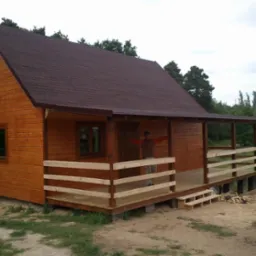 Gotowy, drewniany domek letniskowy z brązowym dachem bitumicznym, otoczony zielenią, z widoczną osobą na werandzie i częściowo widocznym samochodem firmy z danymi kontaktowymi.