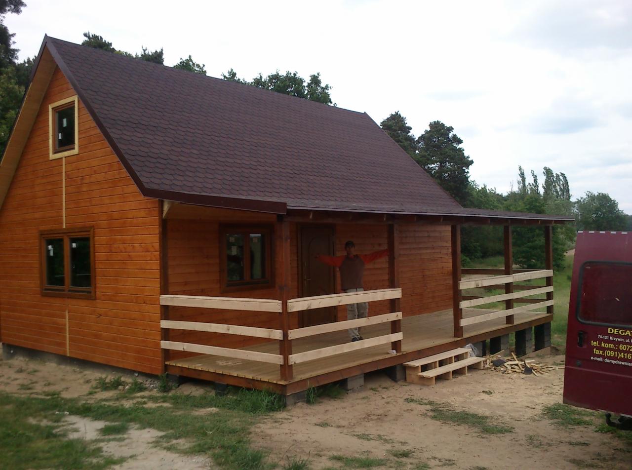 Gotowy, drewniany domek letniskowy z brązowym dachem bitumicznym, otoczony zielenią, z widoczną osobą na werandzie i częściowo widocznym samochodem firmy z danymi kontaktowymi.