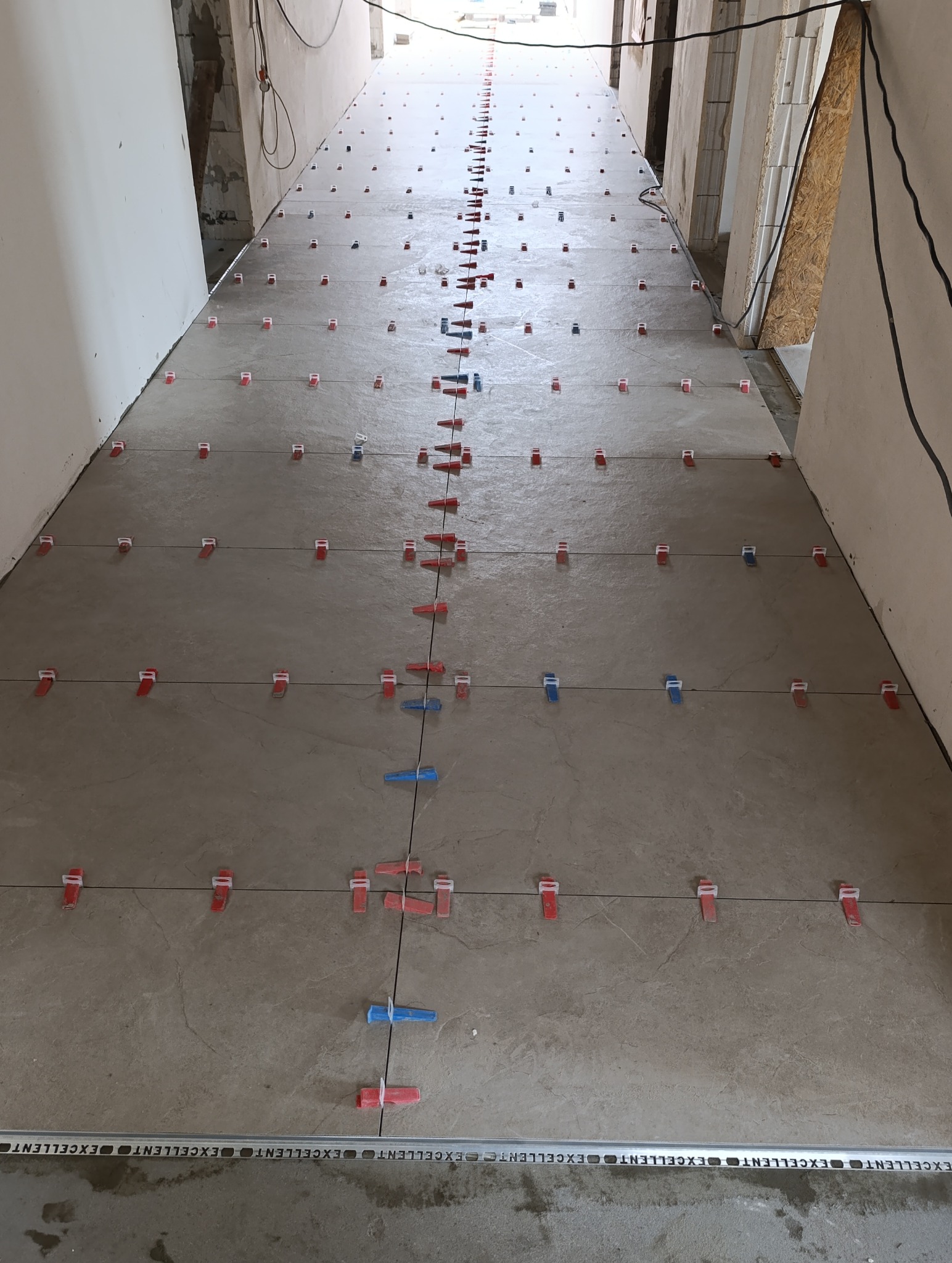 Ułożone płytki podłogowe w korytarzu, z użyciem systemu poziomowania płytek (czerwone i niebieskie klipsy). Widok z góry, perspektywa zbieżna.