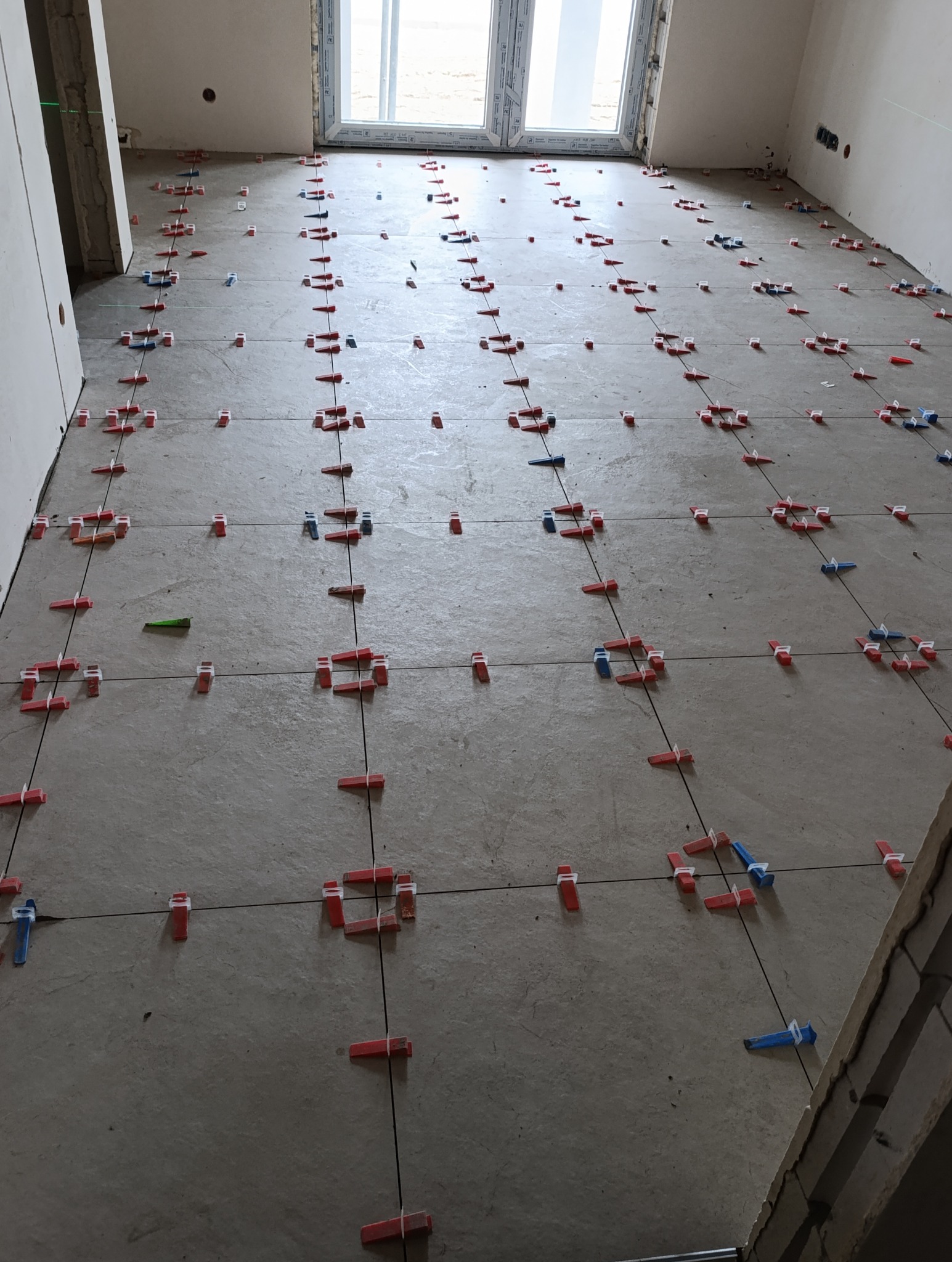 Poziomowanie płytek podłogowych z użyciem klipsów i klinów w jasnym pomieszczeniu z oknem. Widoczne linie wyznaczające siatkę. Etap prac przed fugowaniem.