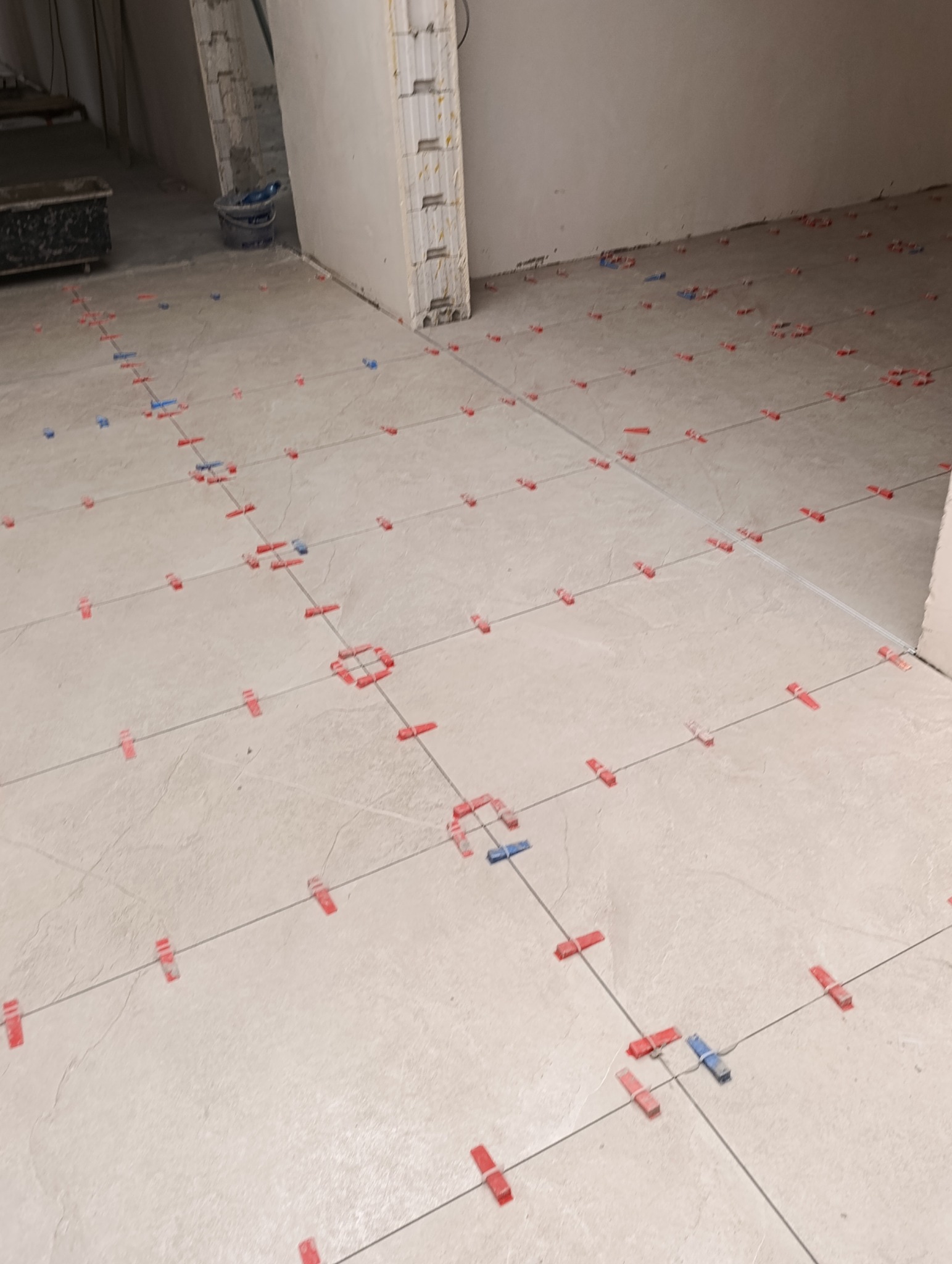 Jasne płytki podłogowe w trakcie układania, z czerwonymi i niebieskimi krzyżykami dystansowymi, widoczne sznurki wyznaczające linie. W tle fragment ściany i wiadro.