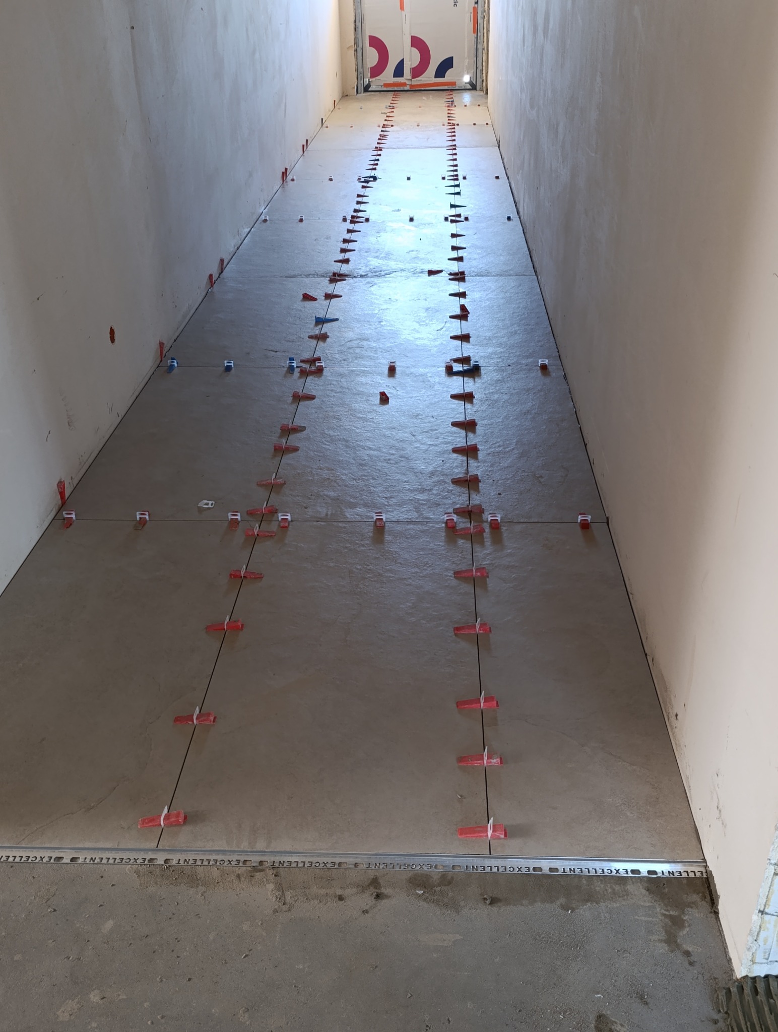 Układanie beżowych płytek podłogowych w korytarzu z użyciem systemu poziomowania płytek (klipsy i kliny). Widoczne fugi i listwa przypodłogowa.