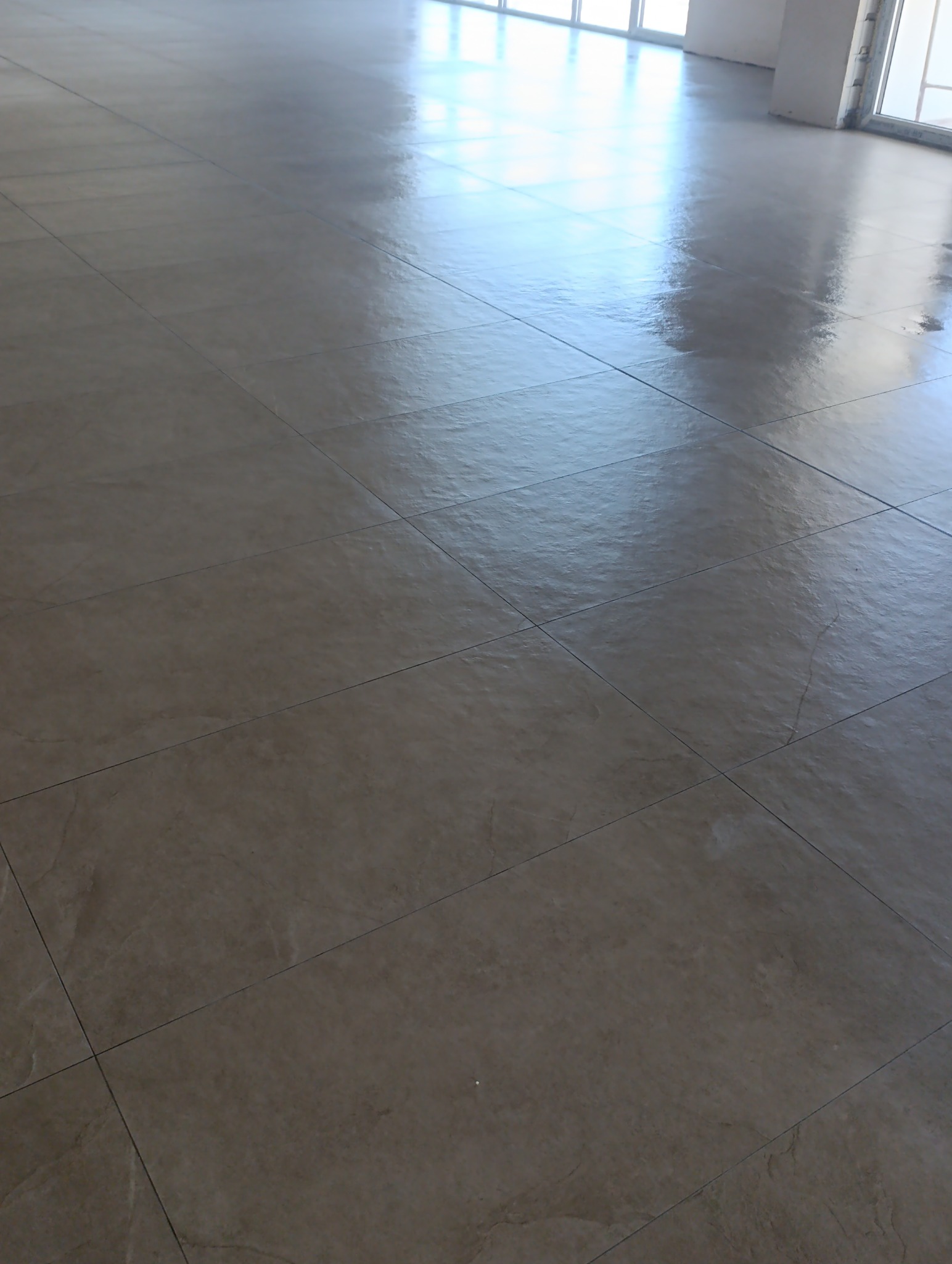 Beżowe, błyszczące płytki podłogowe ułożone w regularny wzór, z widocznym odbiciem światła z okien w tle. Minimalistyczny design, świeżo po realizacji.