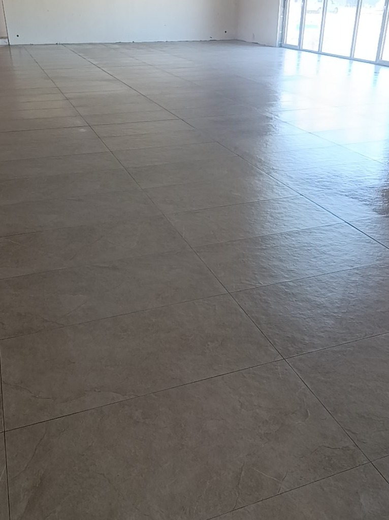 Ułożona, błyszcząca podłoga z szarych, kwadratowych płytek w przestronnym, jasnym pomieszczeniu z dużymi oknami. Widoczna perspektywa i odbicia światła.