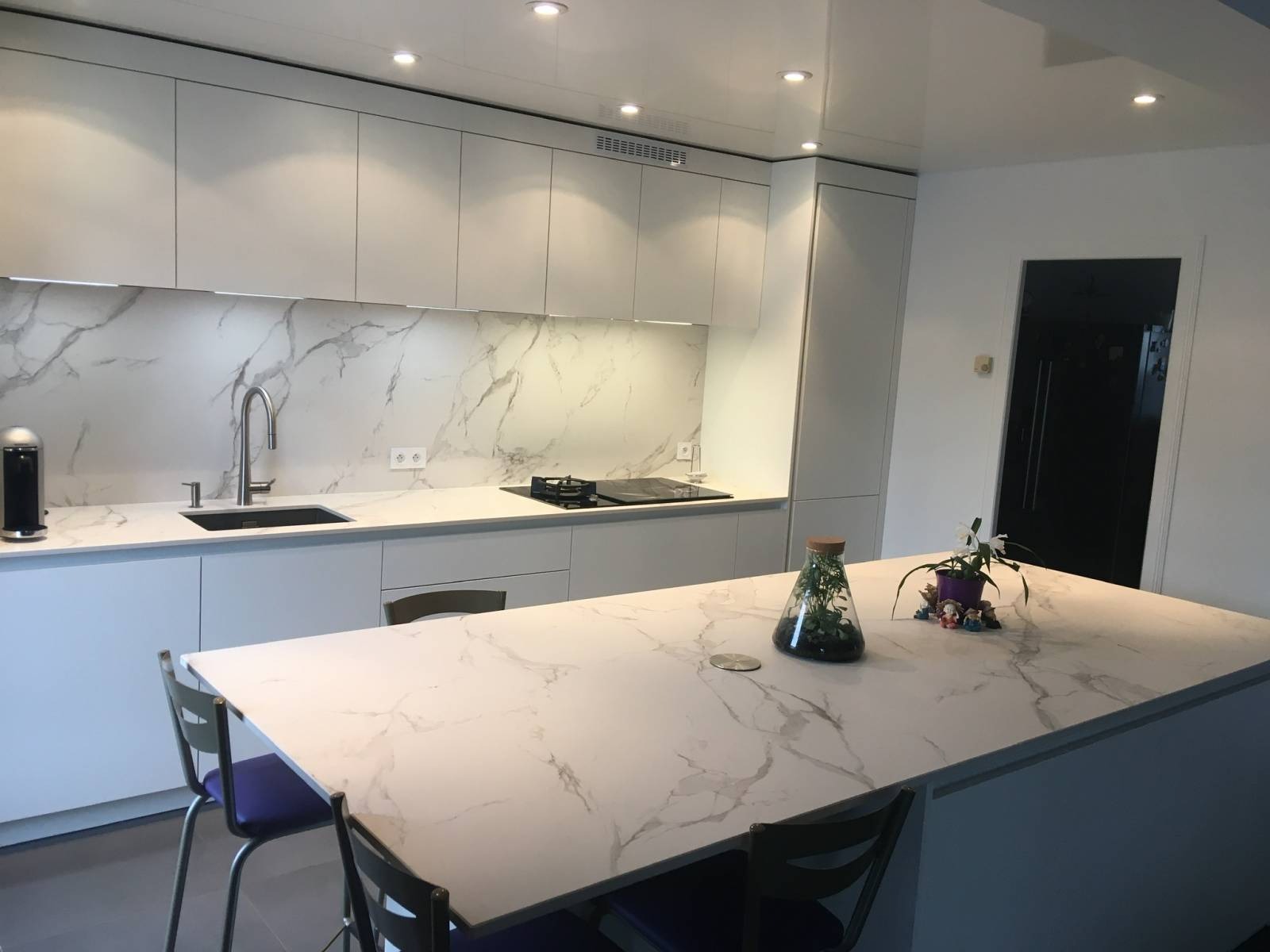 Nowoczesna kuchnia z wyspą i marmurowym blatem. Białe szafki, minimalistyczny design, oświetlenie LED pod szafkami. Na wyspie dekoracje roślinne.