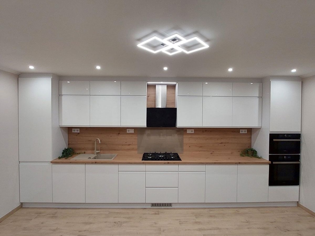 Biała, nowoczesna kuchnia z drewnianym blatem i oświetleniem sufitowym w kształcie kwadratów. Widoczne szafki wiszące i stojące, piekarnik oraz płyta gazowa.