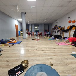 Tu I teraz - Grupa osób w pozycji relaksacyjnej na matach do jogi w przestronnym, jasnym pomieszczeniu z dekoracjami halloweenowymi w tle.