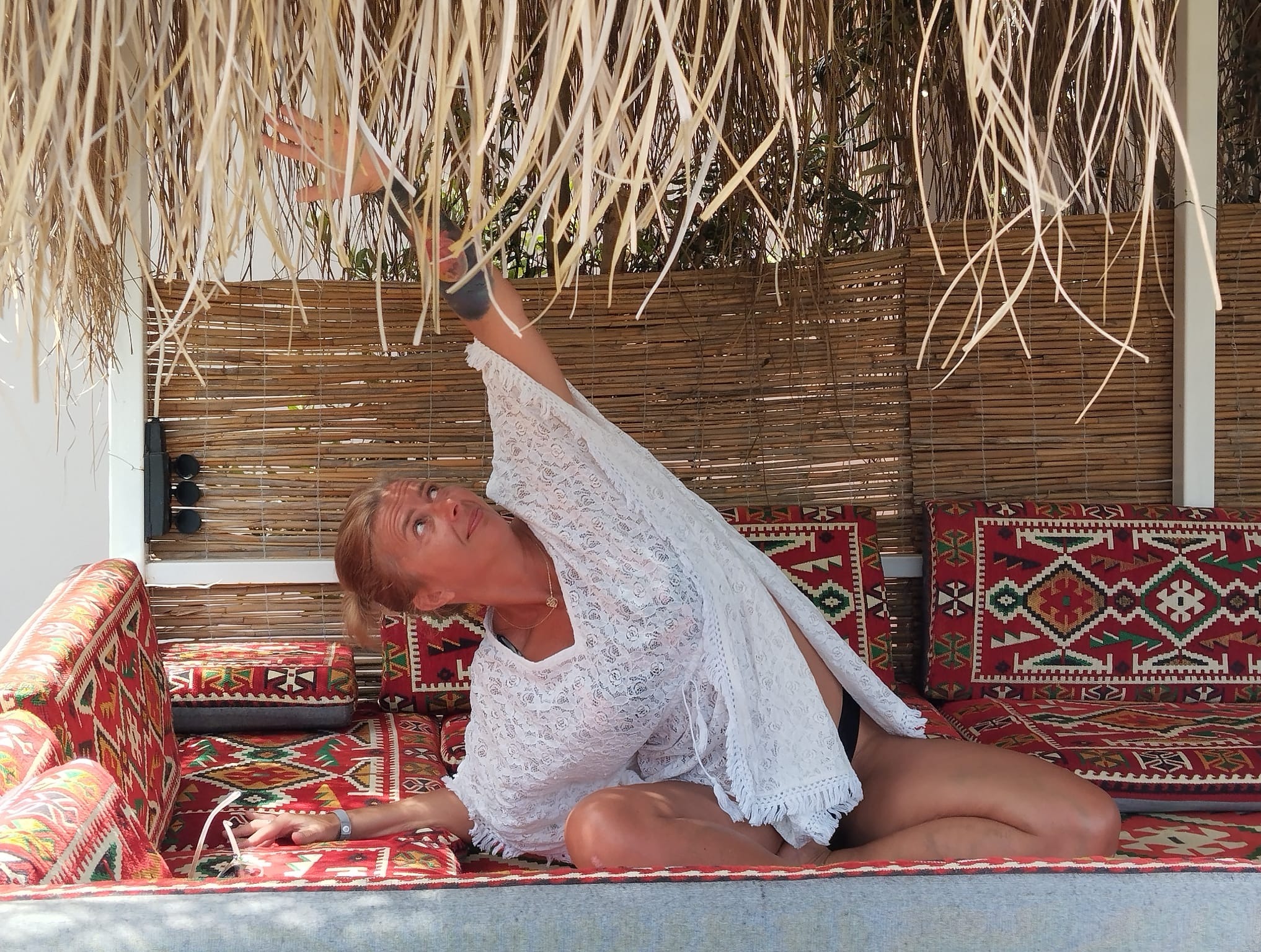 Kobieta w białej tunice wykonuje pozycję jogi na wzorzystej sofie pod strzechą. Widok z dołu, skupienie na twarzy i wyciągniętej ręce.
